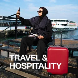 Person in black outfit sitting on a dock taking a selfie with a red suitcase nearby and boats in the background.