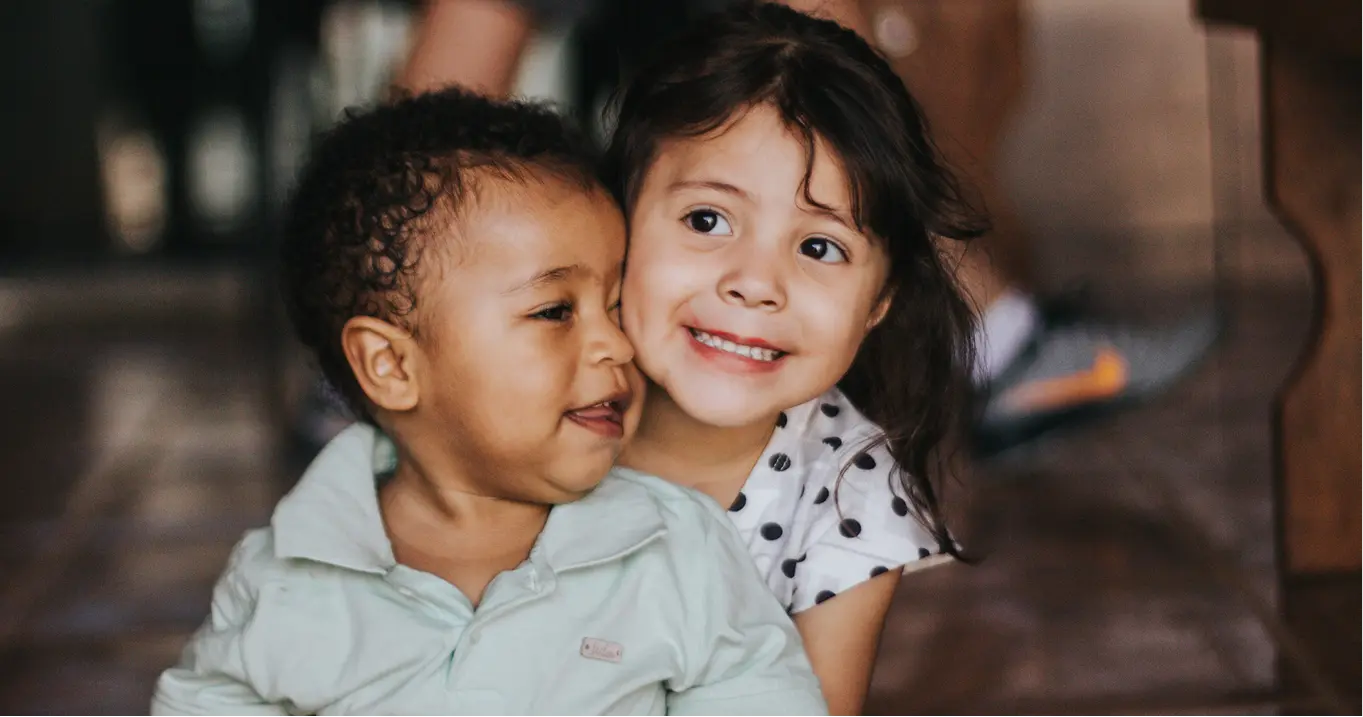 Two young children smiling and hugging indoors with blurred background.