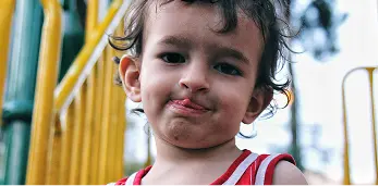 Young child wearing a red and white striped tank top making a playful face outdoors near playground equipment.