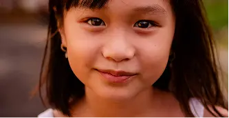 Close-up of a smiling young girl with dark hair and brown eyes outdoors.