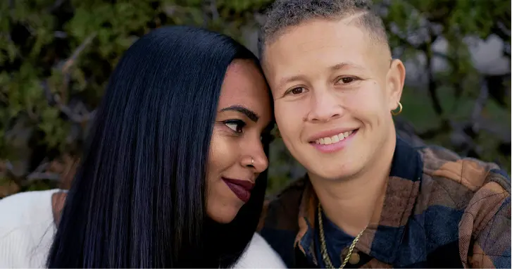 Two people smiling closely together outdoors with greenery in the background.