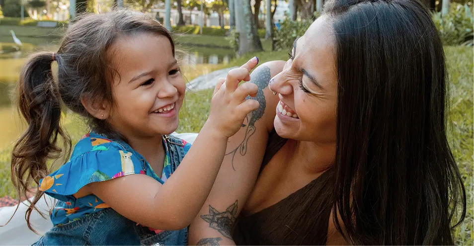 Smiling young girl playfully touching the nose of a laughing woman with tattoos, outdoors near greenery and water.