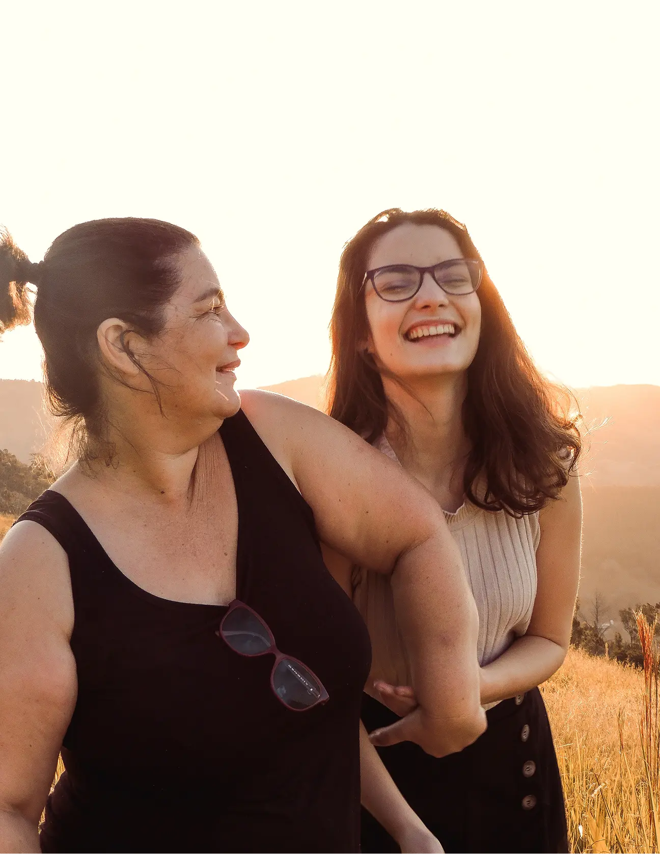 Two women outdoors at sunset, one with glasses laughing and linking arms with the other smiling woman.