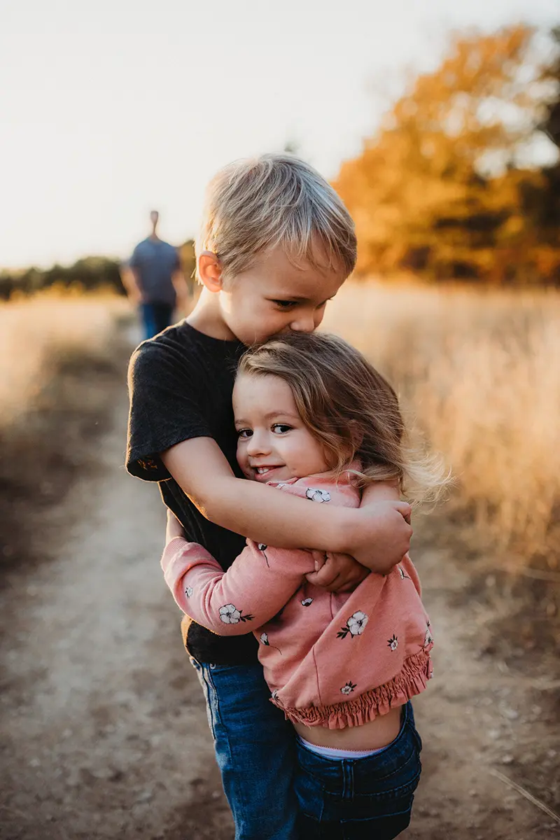 Young boy hugging and kissing a smiling girl outdoors on a dirt path during golden hour.