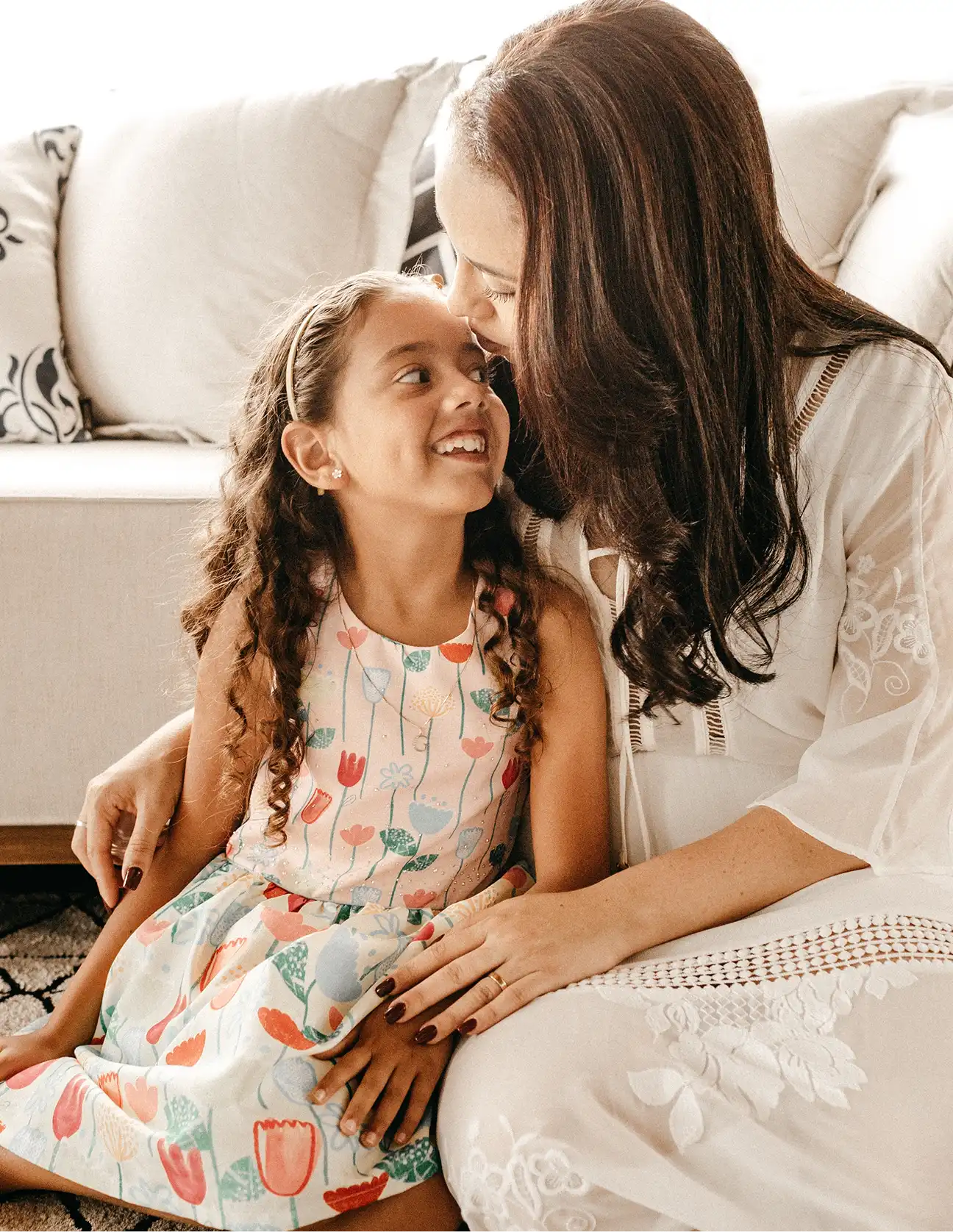 A mother lovingly kisses her smiling daughter on the forehead as they sit together on a couch.