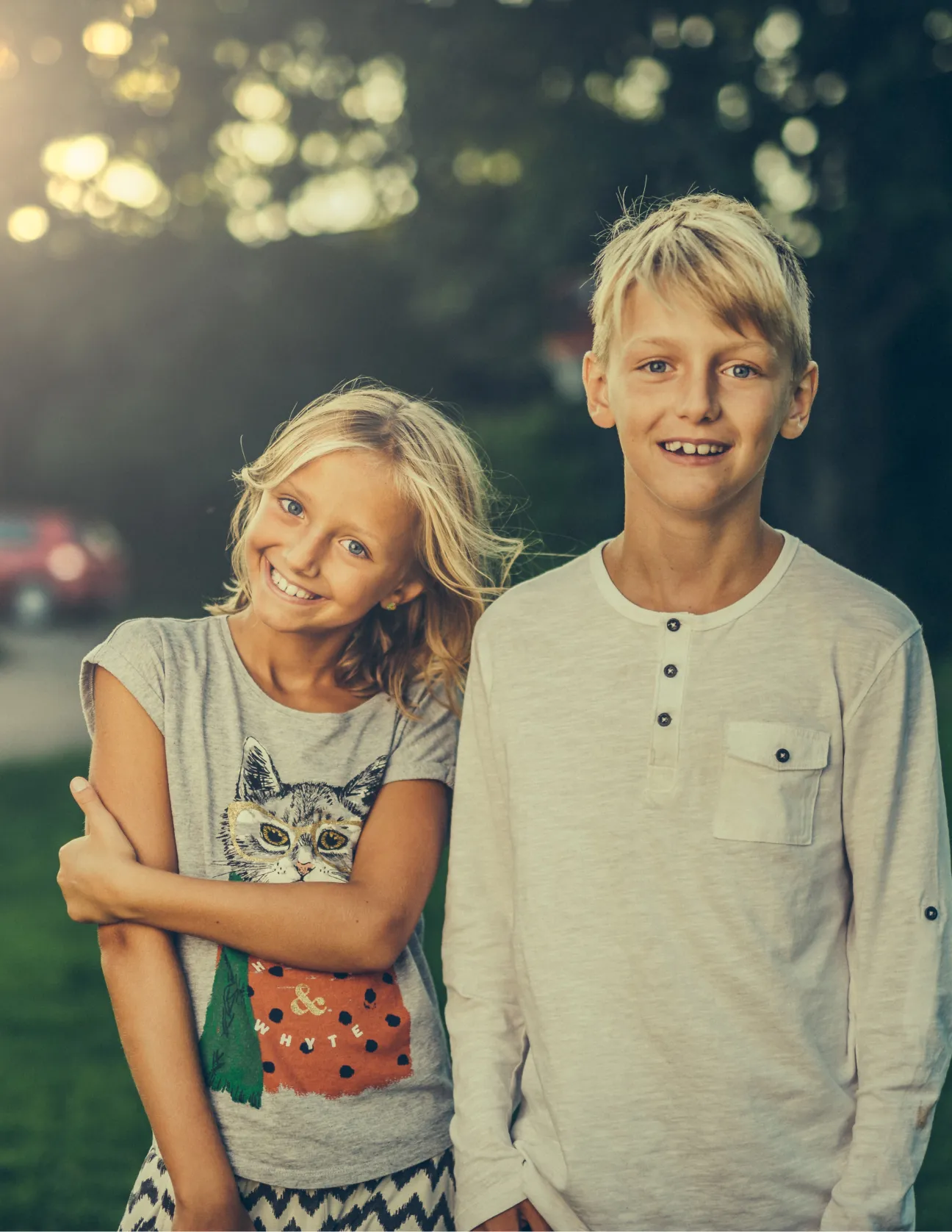Smiling boy and girl standing outdoors in soft sunlight, with a blurred natural background.