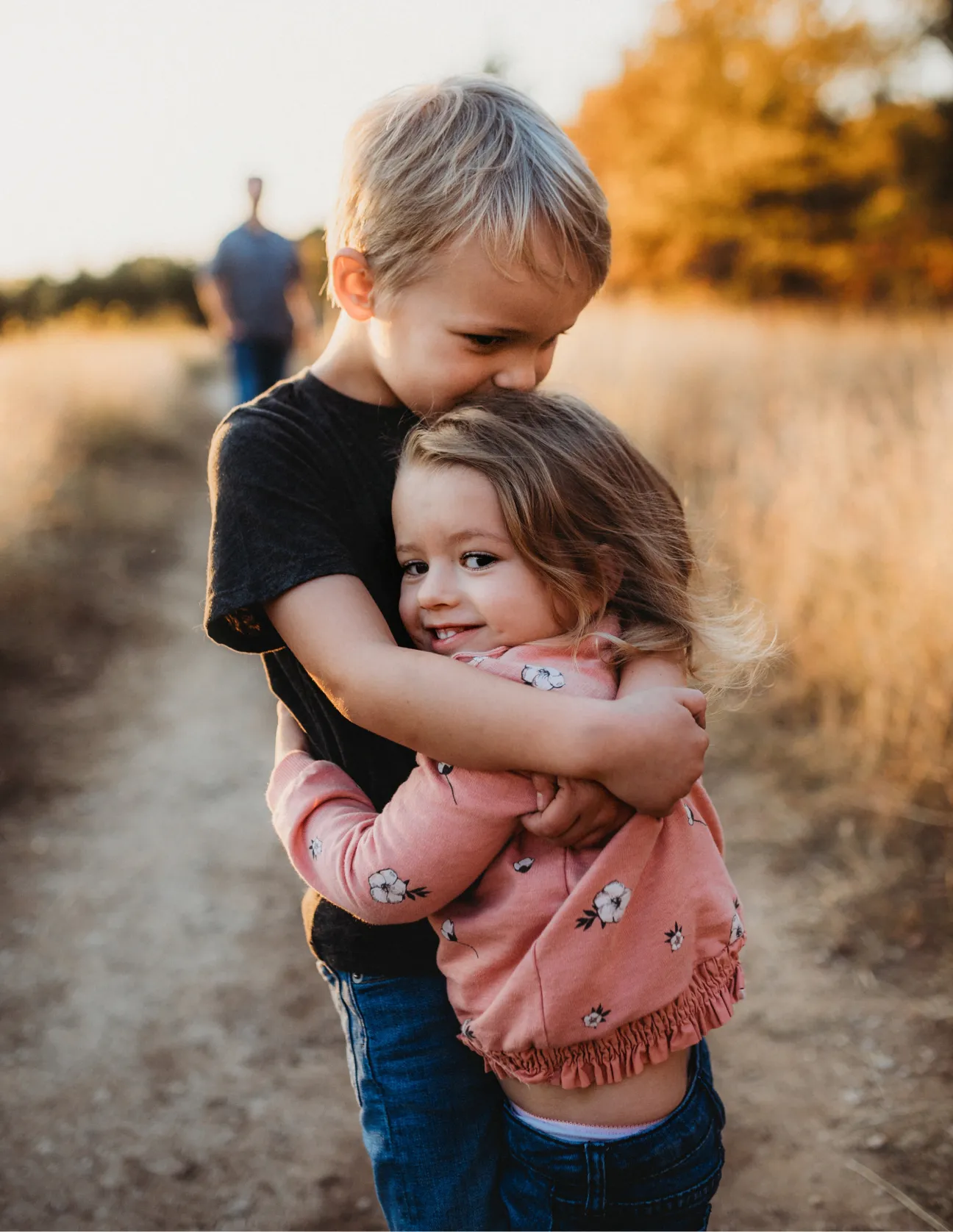 A young boy hugging and kissing the head of a smiling little girl outdoors on a dirt path with golden foliage in the background.