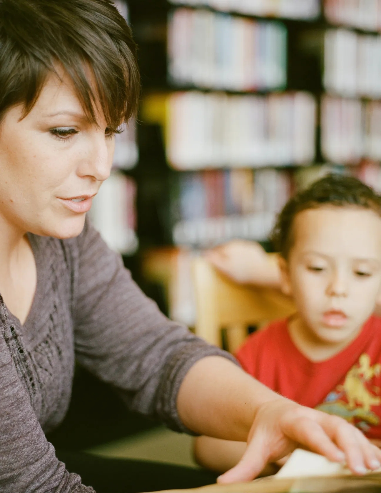 Woman reading a book to a young boy in a library setting with blurred bookshelves in the background.