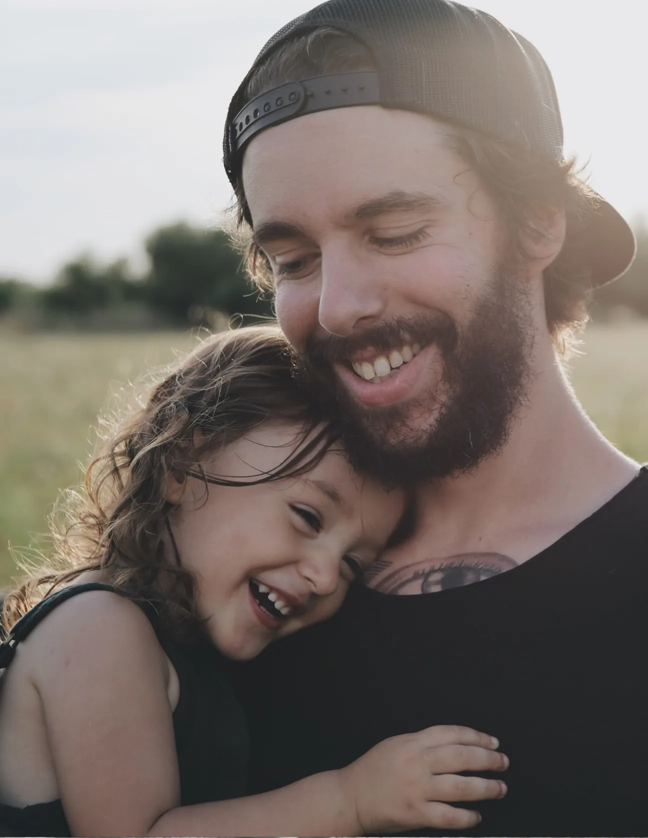 Smiling bearded man wearing a backward cap hugs a laughing young girl outdoors at sunset.