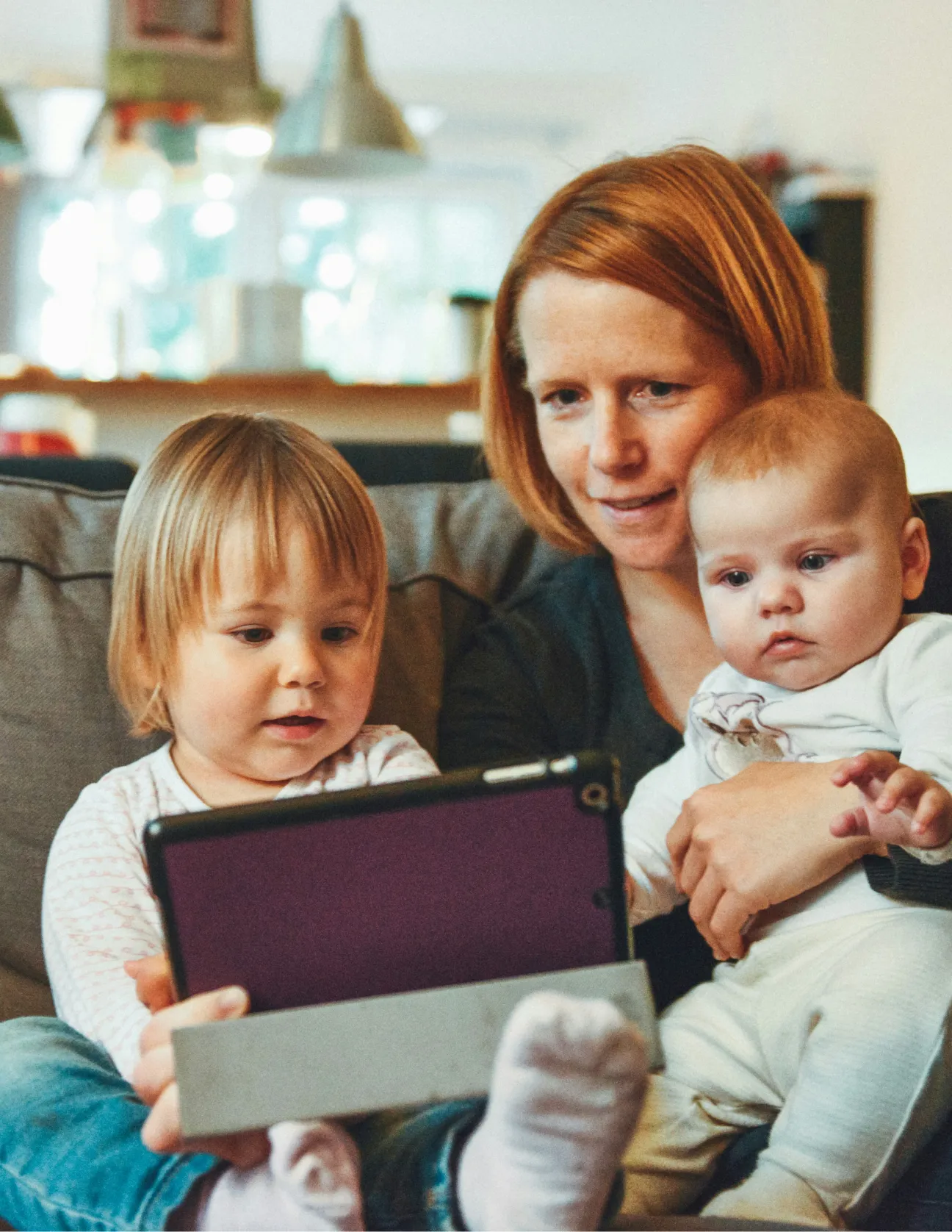 Mother sitting on a couch holding a baby, with a toddler watching a tablet in front of them.