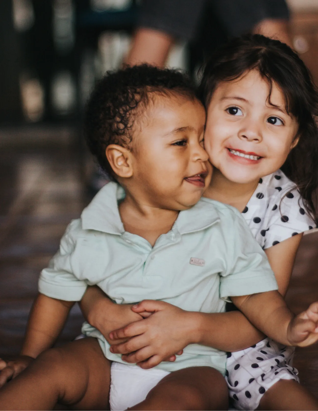 Smiling young girl hugging a baby boy who is wearing a light green shirt and diaper.