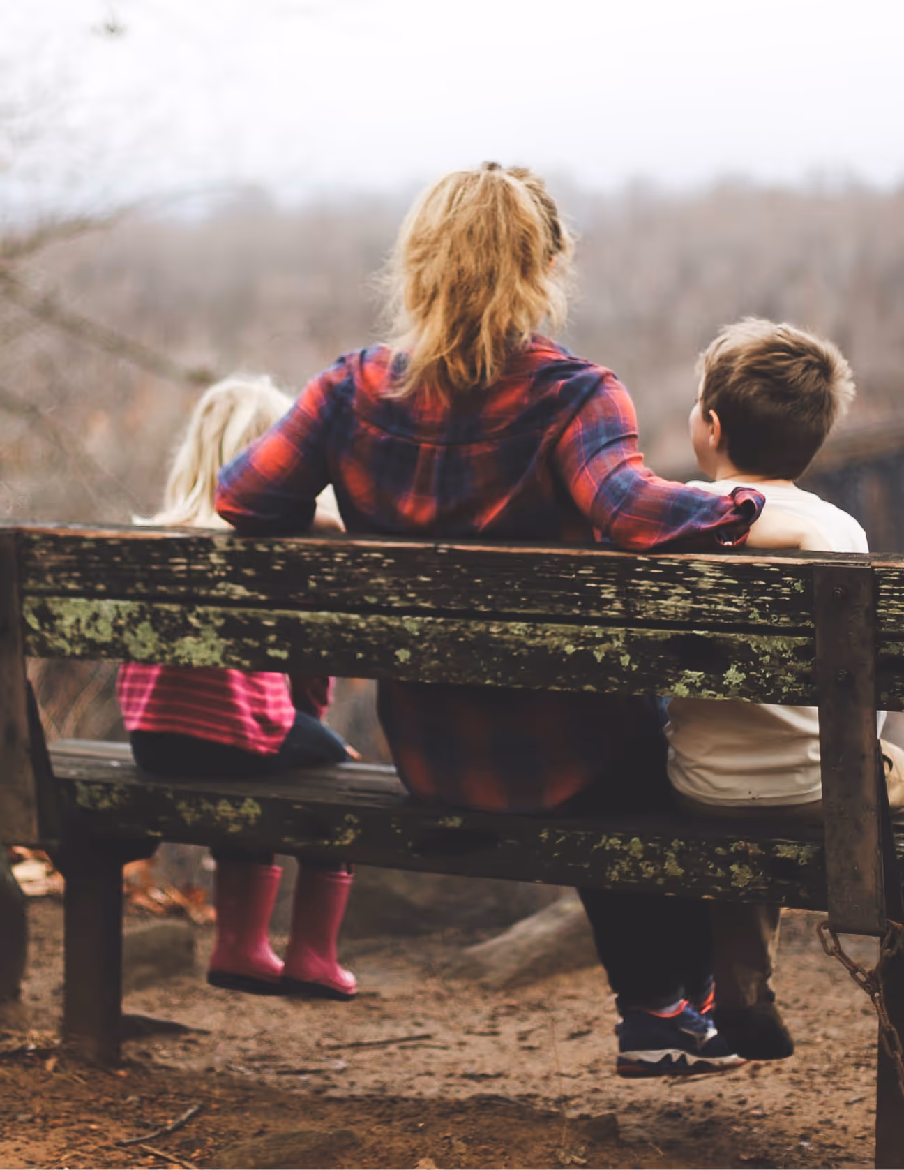 Rear view of a woman and two children sitting on a weathered wooden bench outdoors, with the woman’s arm around the boy.