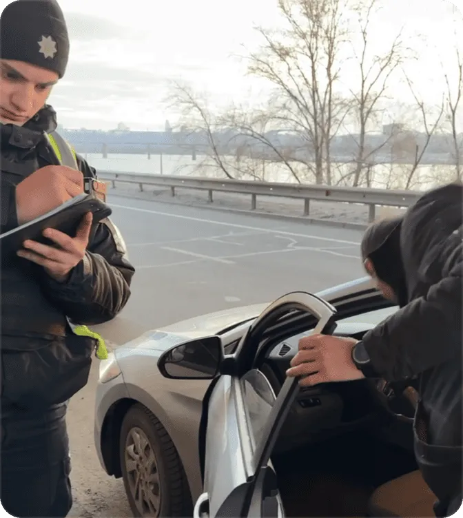 Police officer writing on a notepad while a man leans into the open door of a silver car on the roadside.