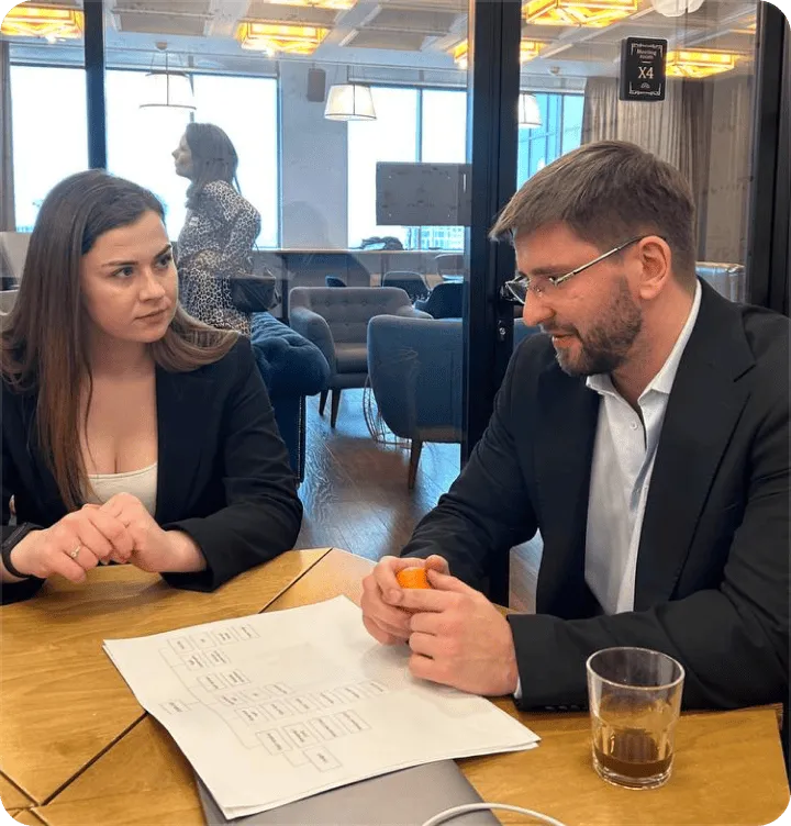 Man and woman in business attire having a discussion at a wooden table with papers and a laptop in an office setting.