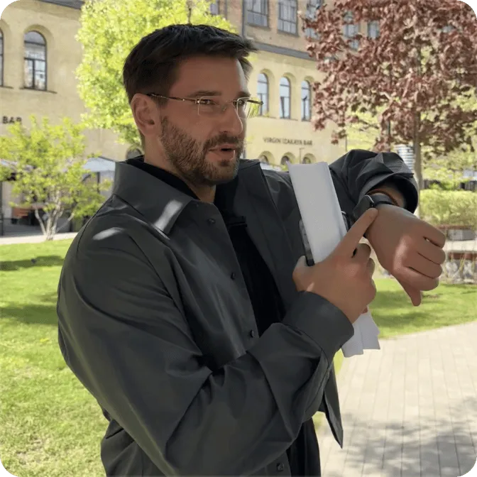 Man in dark jacket looking at and pointing to a smartwatch on his wrist while holding papers outdoors in a park.