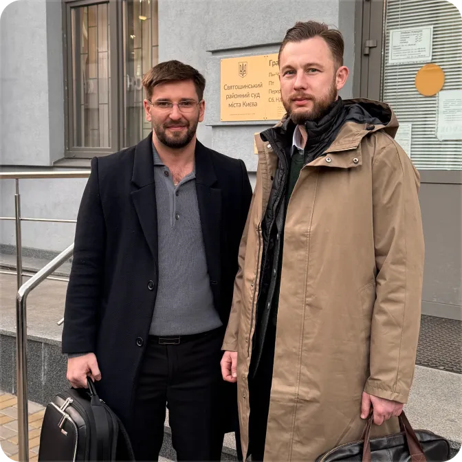 Two men standing outside a building with a plaque, each holding a briefcase and wearing coats.