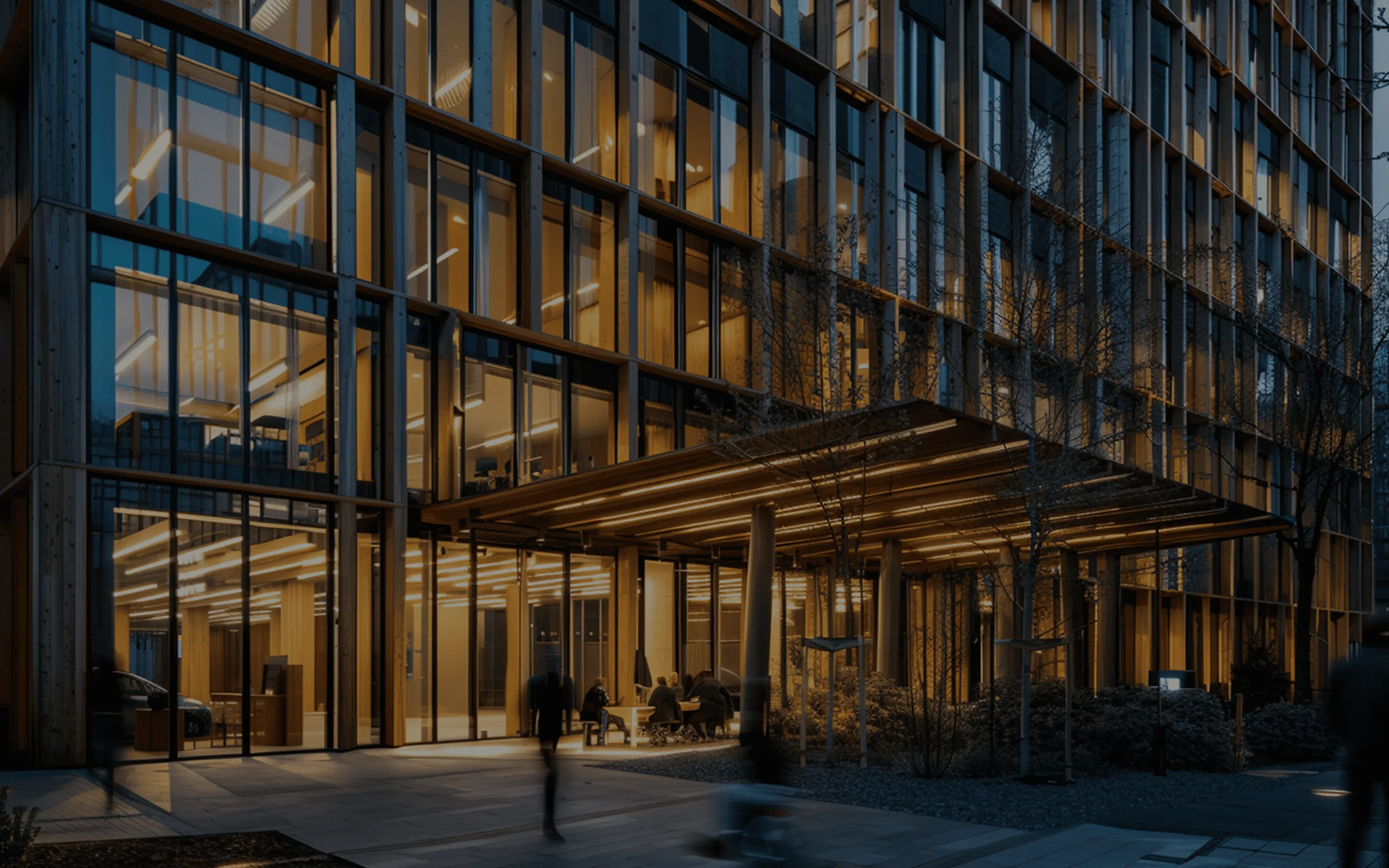 Modern office building entrance illuminated with warm lights at dusk, showing people sitting under a canopy and blurred figures walking by.