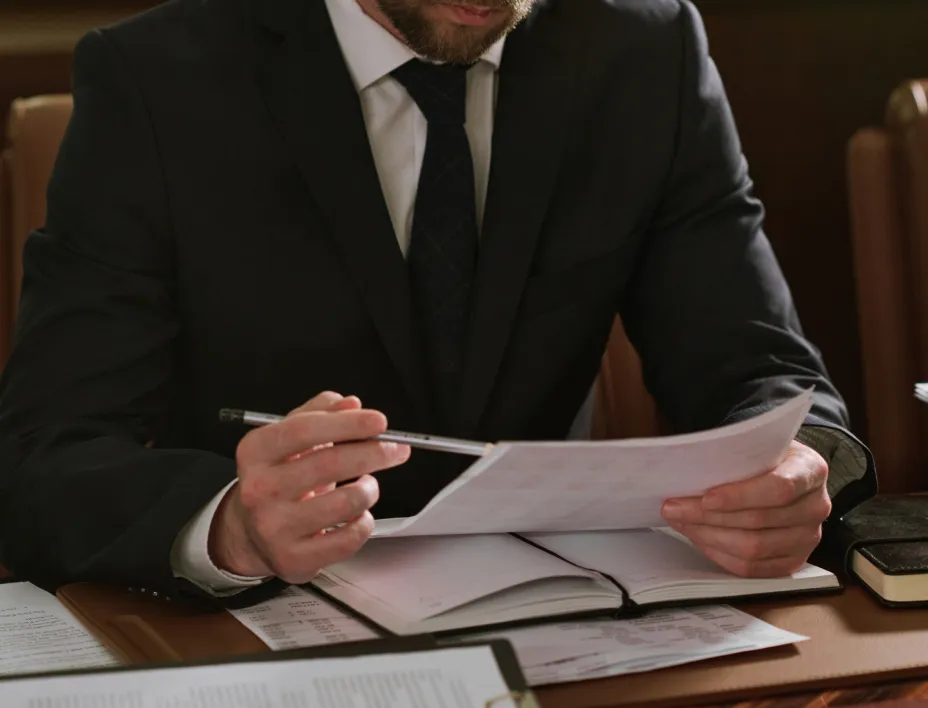 Man in a black suit examining documents with a pen in hand at a desk.