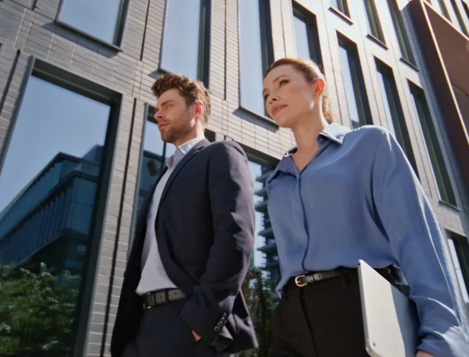 Businessman in a dark suit and businesswoman in blue blouse walking outside a modern office building.