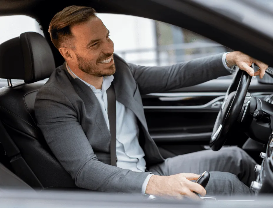 Smiling man in a gray suit driving a car, holding the steering wheel with one hand and the gear shift with the other.
