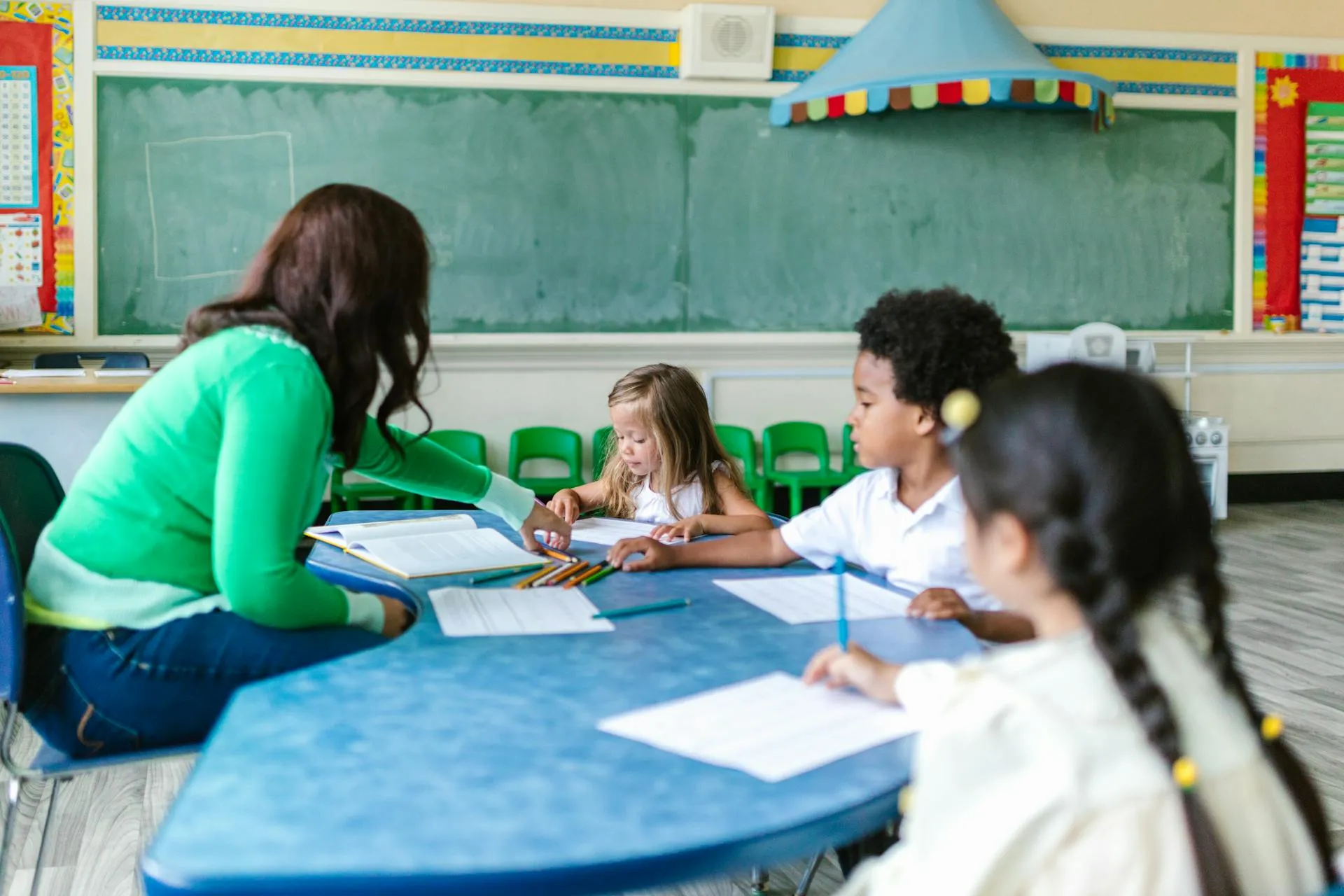 Teacher in green sweater assisting young students sitting at a blue table in a classroom.