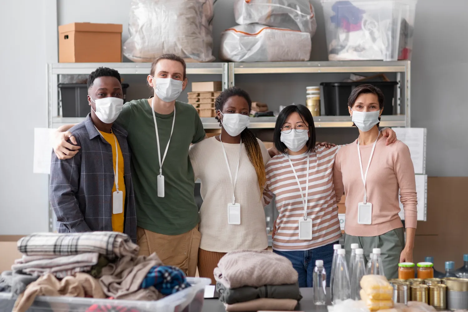 Diverse group of five masked volunteers standing side by side with arms around each other in a storage room with supplies.