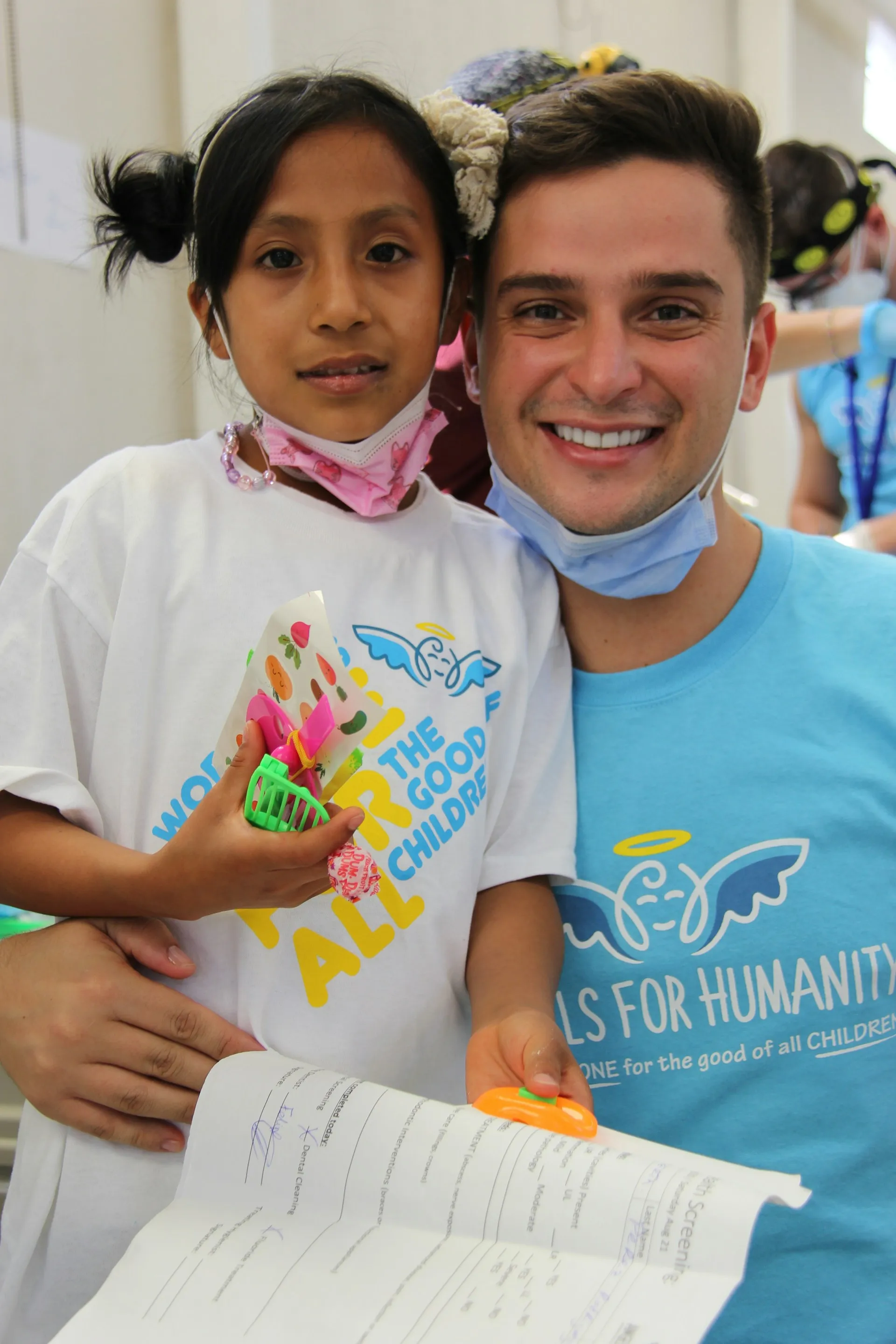 Smiling man and young girl wearing 'Angels for Humanity' T-shirts, holding a document and small toys indoors.