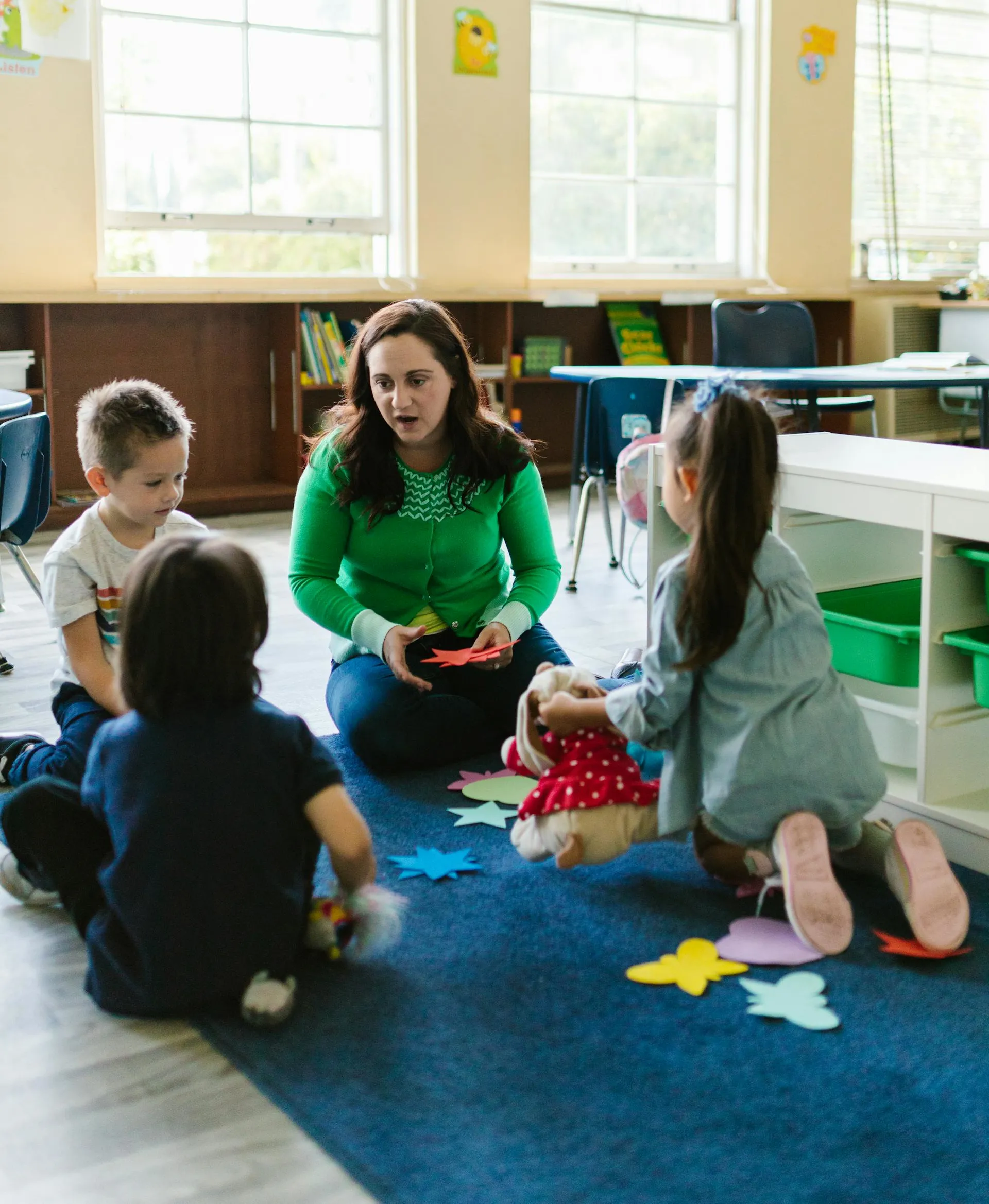 Teacher sitting on classroom floor showing colorful paper shapes to three young children engaged in group activity.