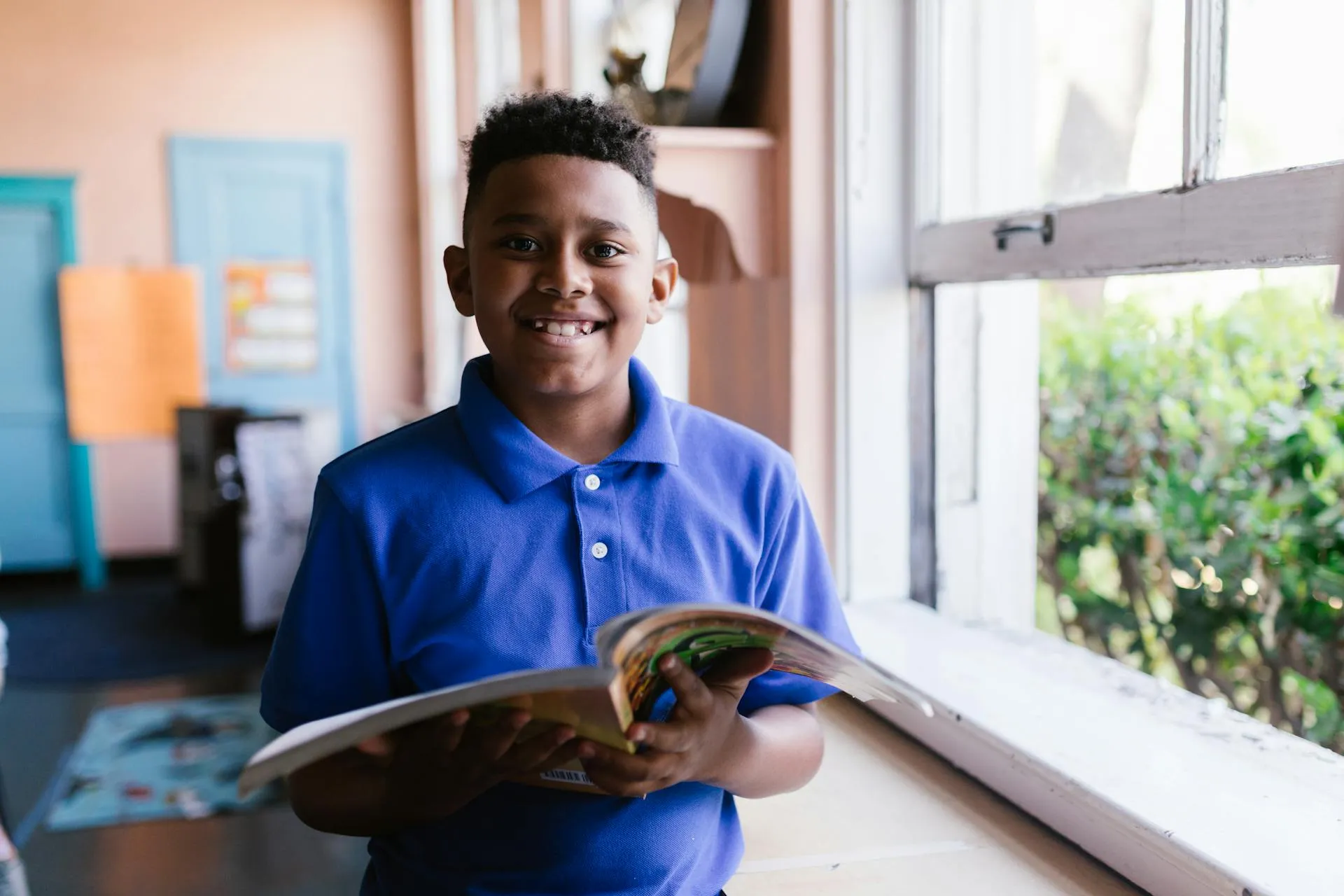 Smiling boy in a blue polo shirt holding an open book by a window with greenery outside.