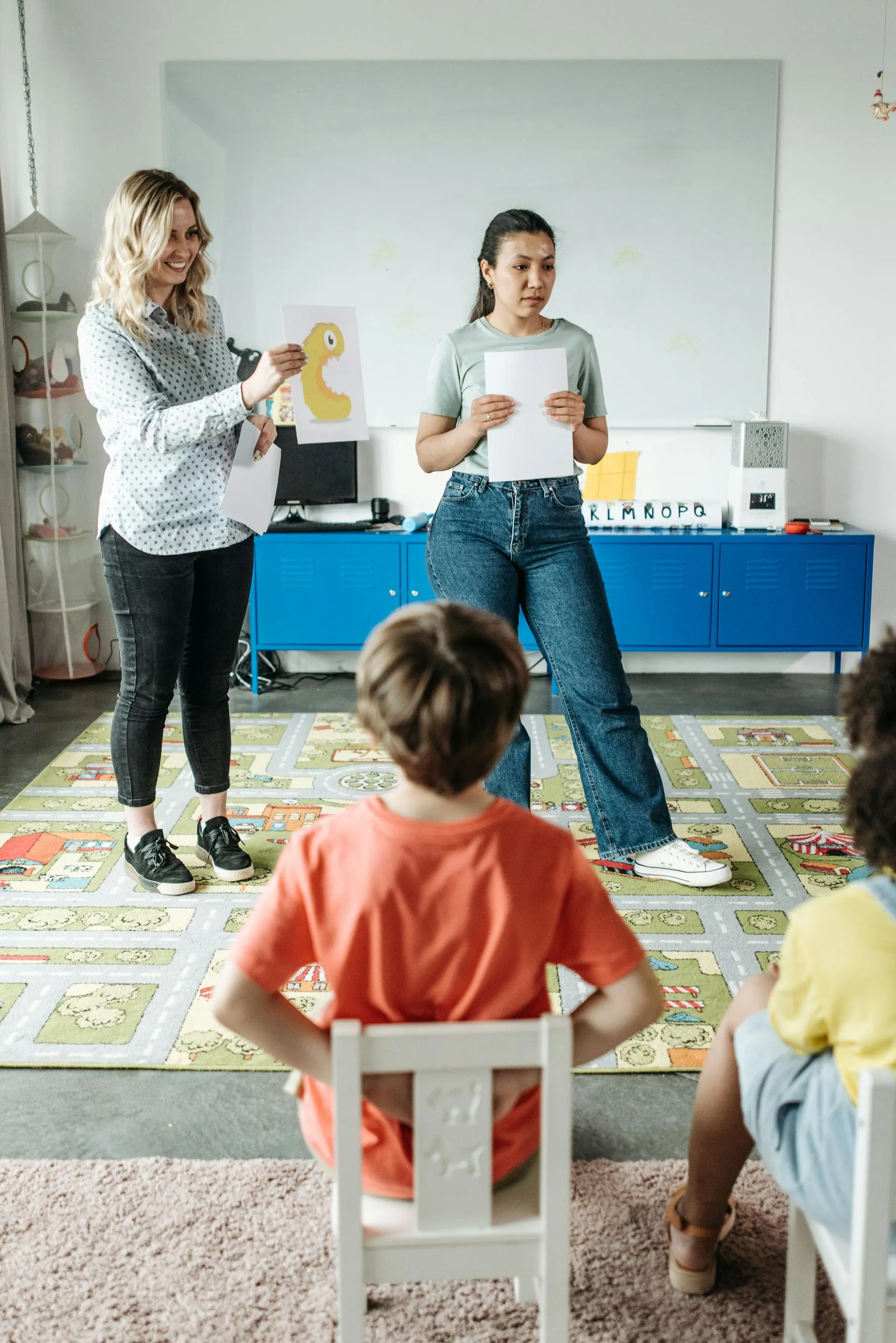 Young girl in a red shirt writing on paper while sitting among other children in a classroom with sunlight coming through window blinds.