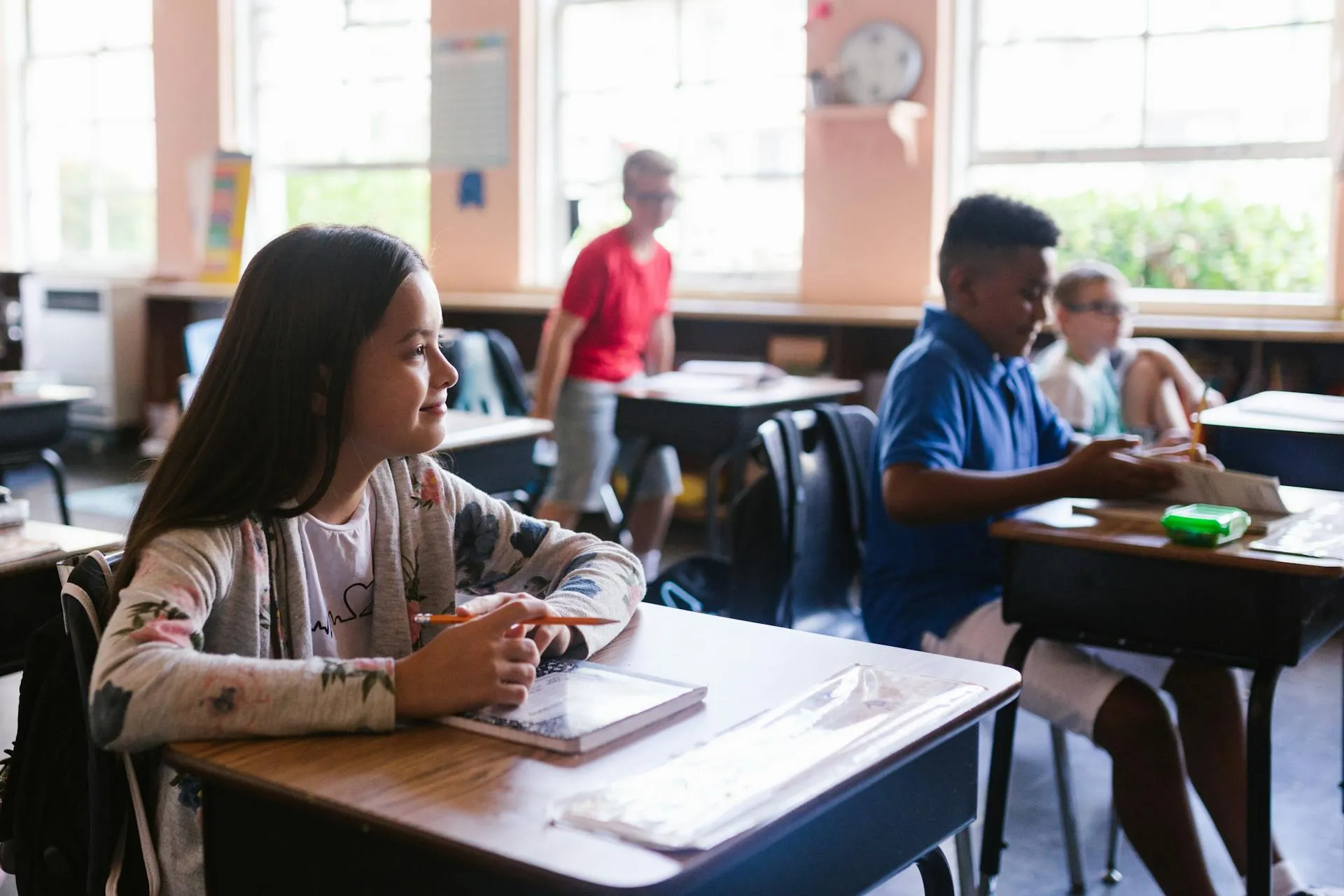 Girl sitting at a classroom desk holding a pencil and looking ahead, with other students and a teacher in the background.