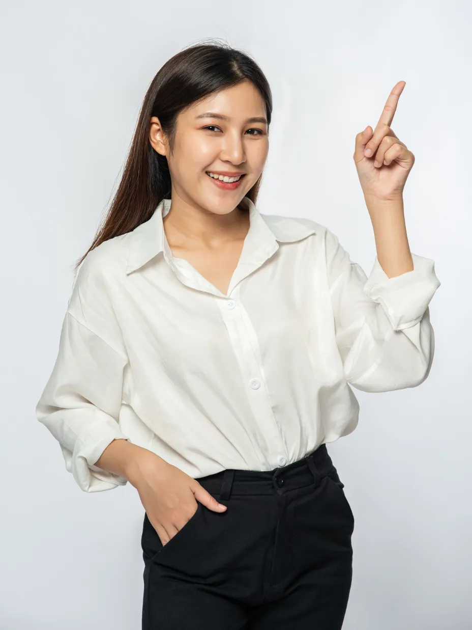 Smiling woman wearing white shirt and black pants pointing upward with her right index finger.