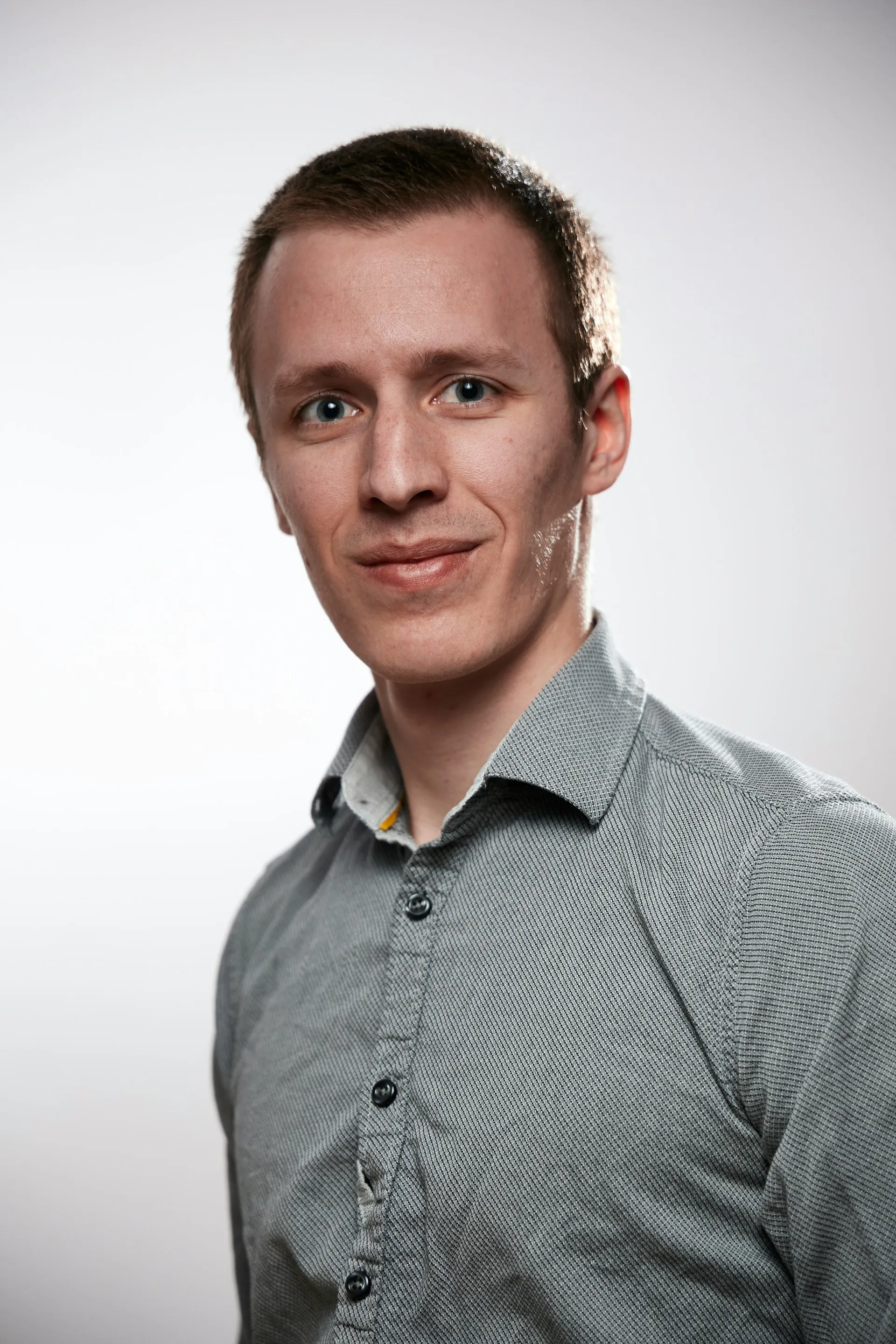 Portrait of a young man with short brown hair wearing a gray button-up shirt against a plain background.