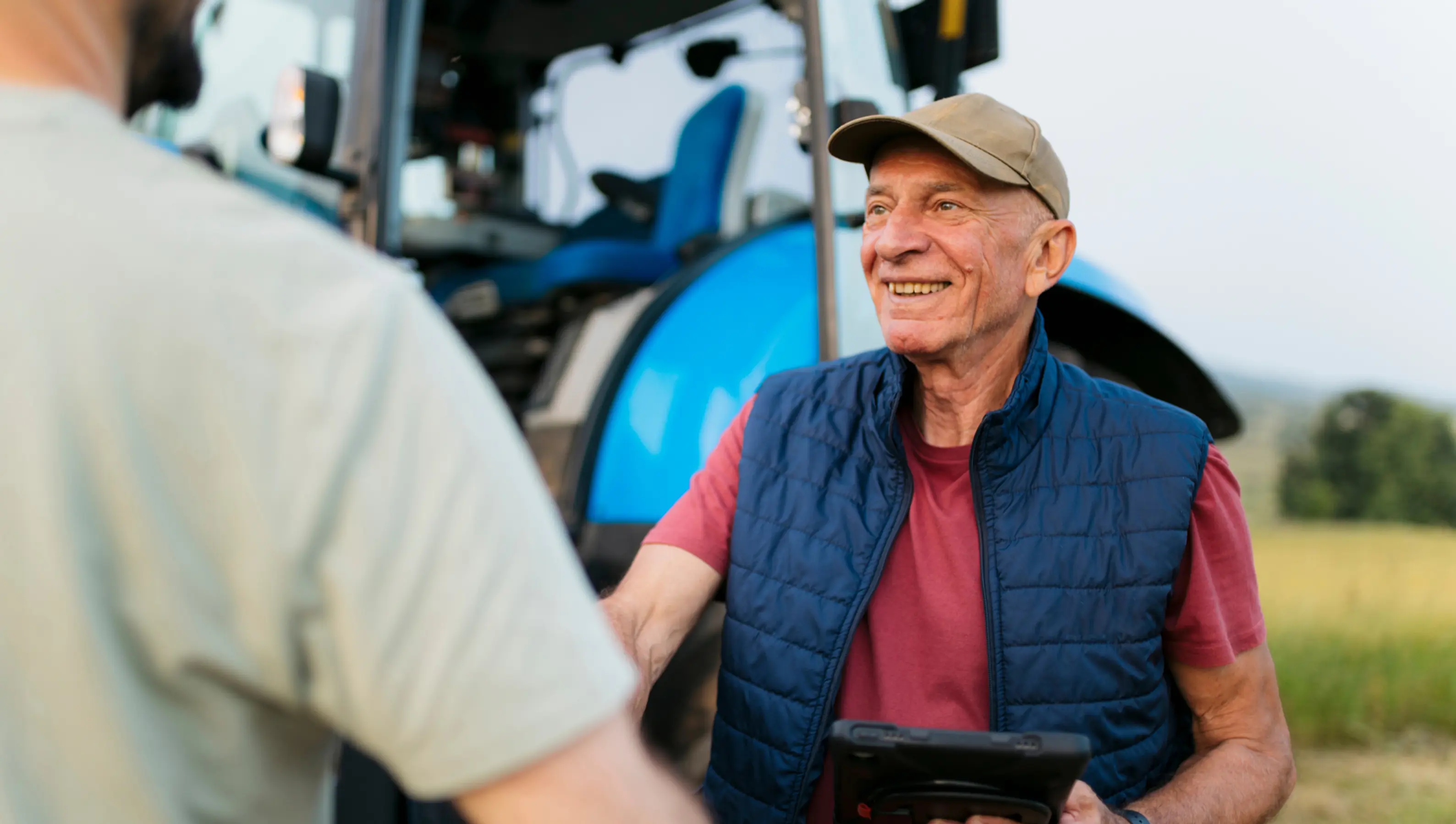 Älterer Landwirt mit Baseballkappe und blauem Steppweste, lächelnd und Tablet haltend, vor blauem Traktor im Freien.
