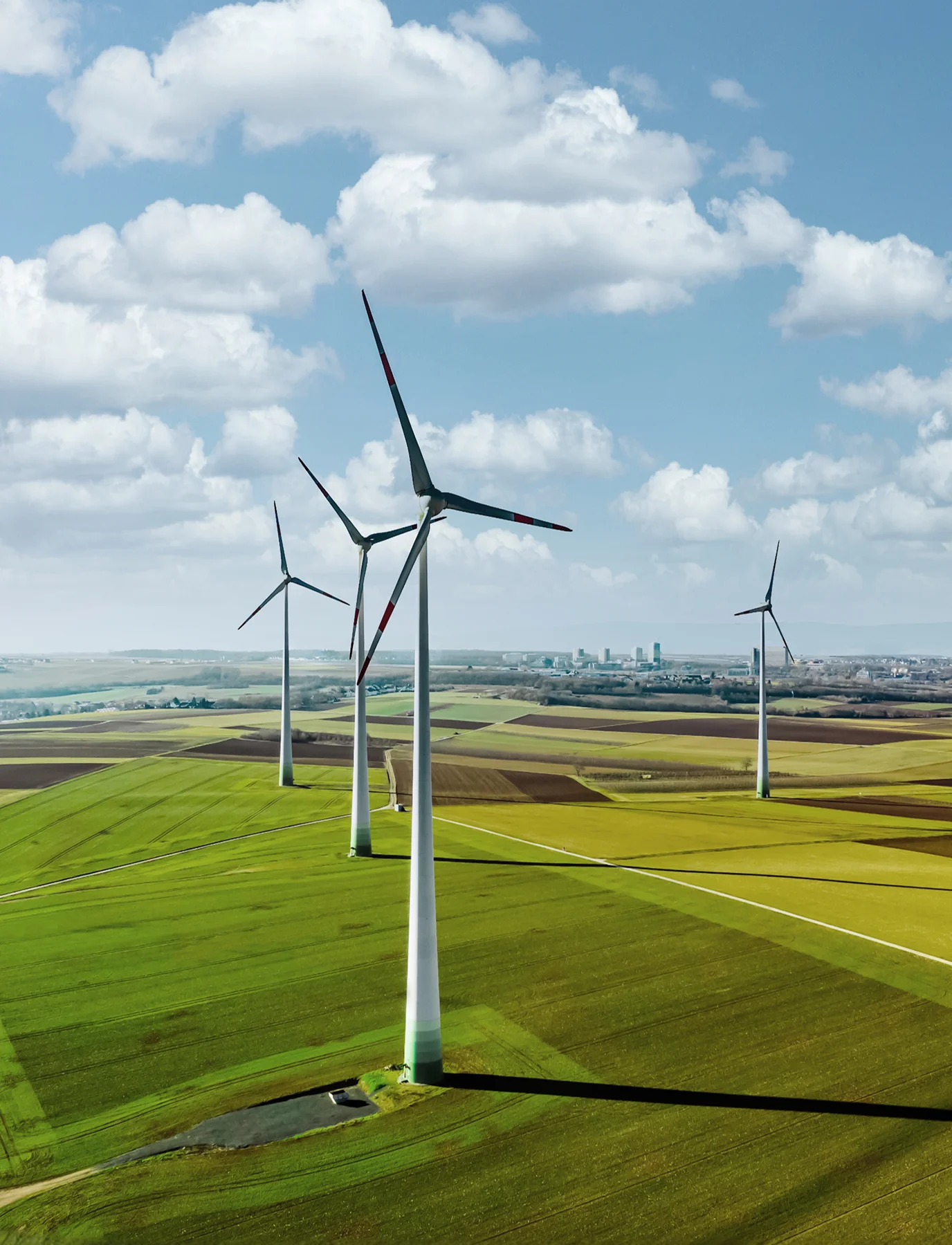 Mehrere Windkraftanlagen auf grünen Feldern unter einem bewölkten Himmel, mit Stadt im Hintergrund.