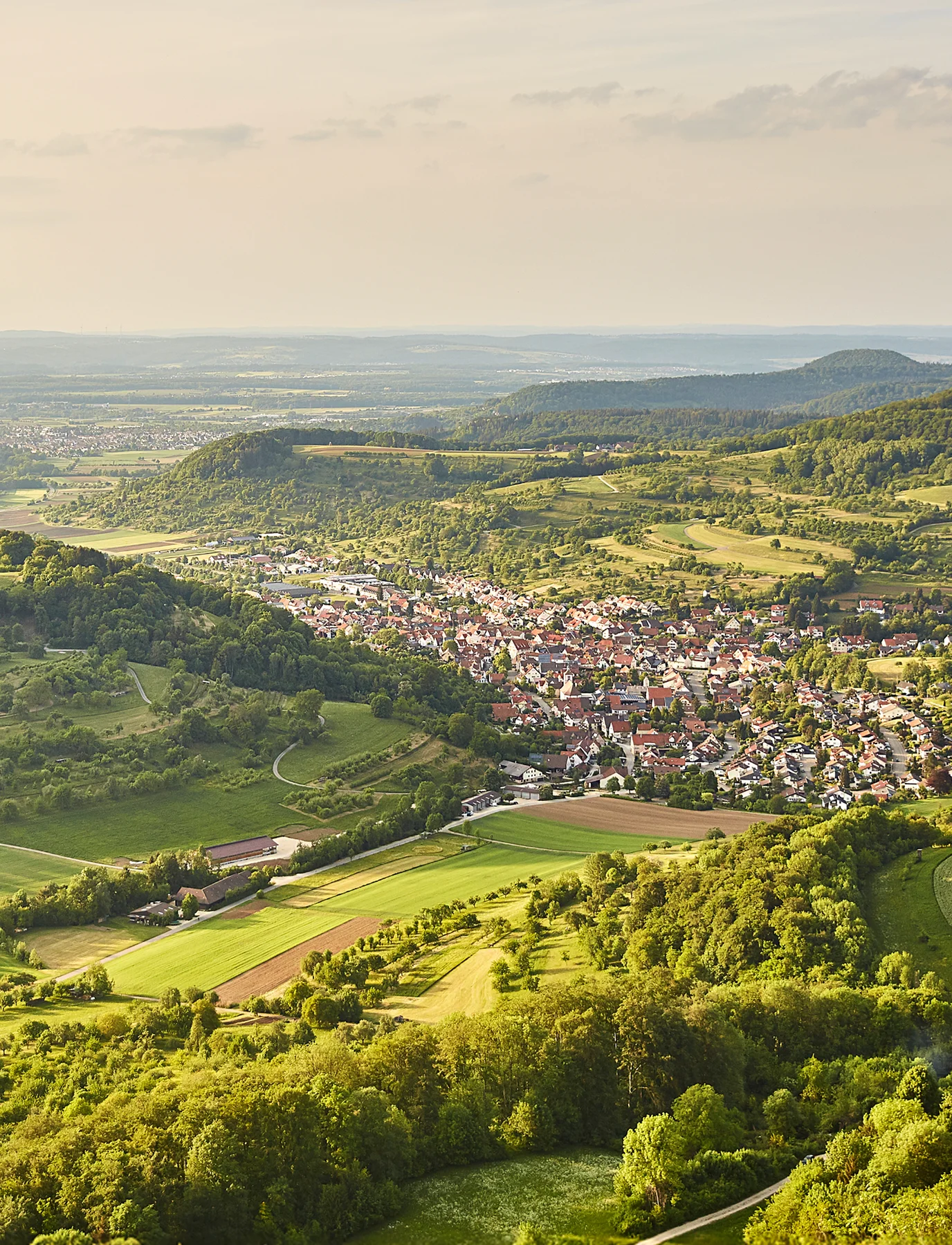 Luftaufnahme eines kleinen Dorfes umgeben von grünen Feldern, bäumen und sanften Hügeln unter einem bewölkten Himmel.
