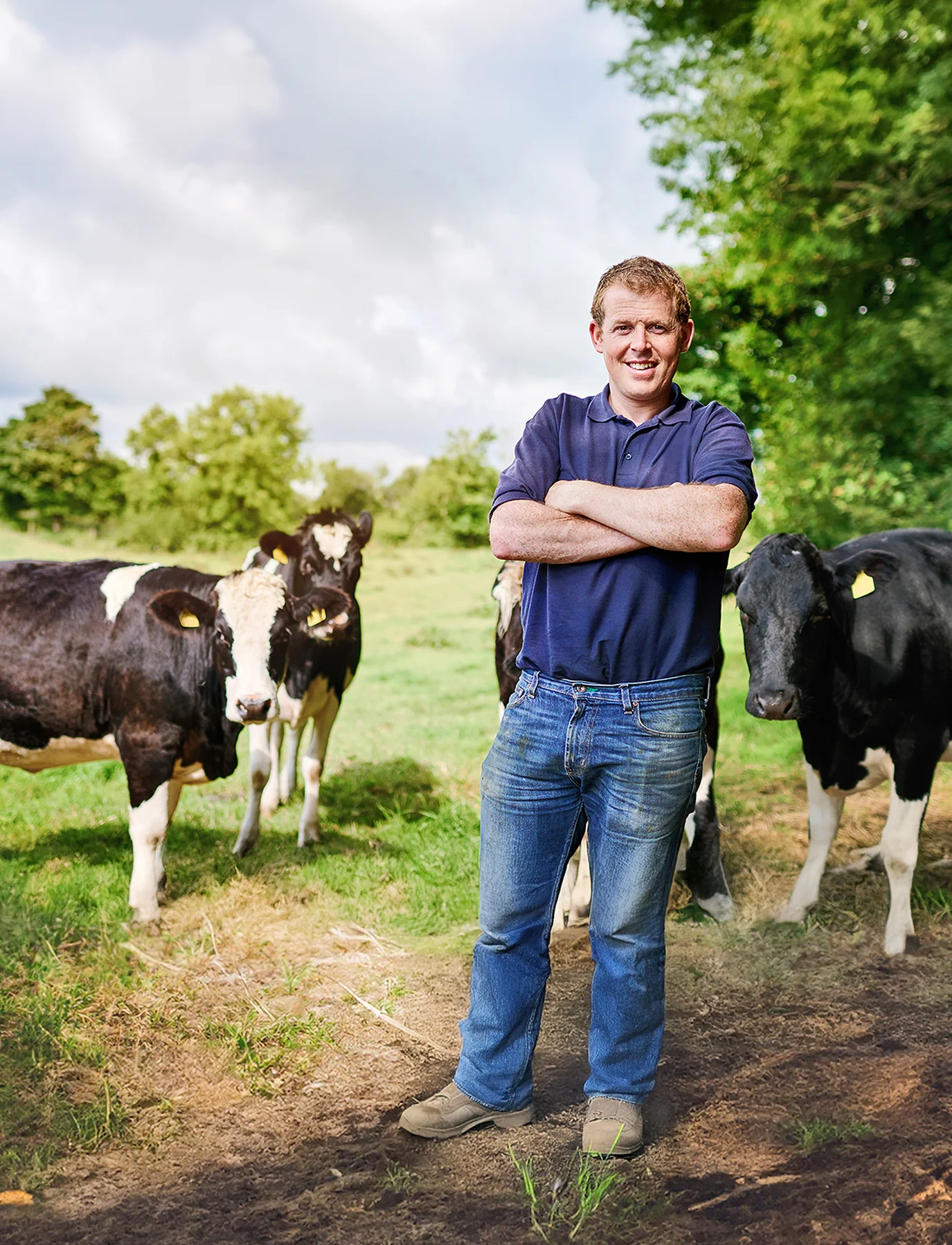 Ein lächelnder Landwirt steht mit verschränkten Armen auf einer Wiese neben mehreren schwarz-weißen Kühen.