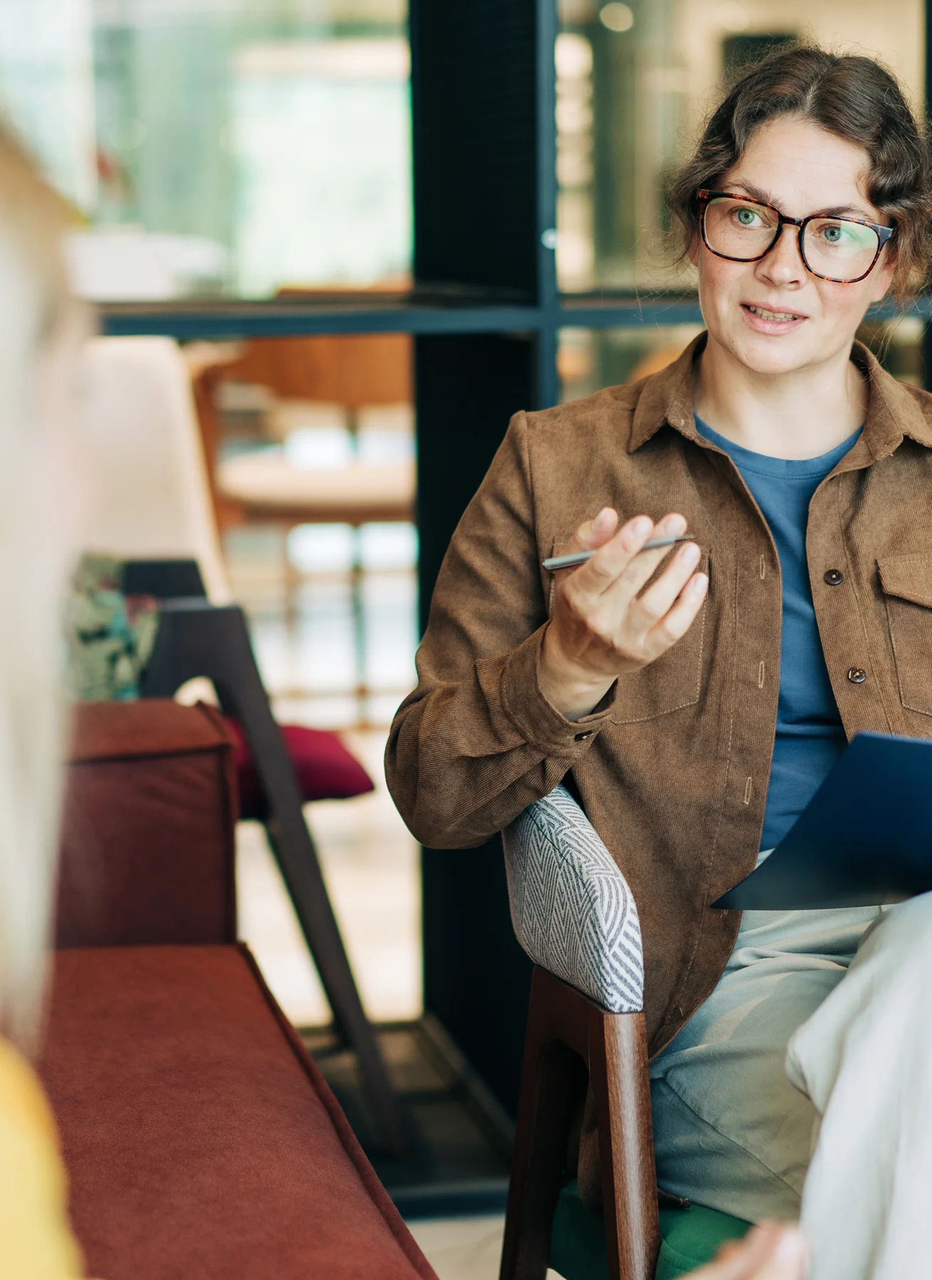 Frau mit Brille und brauner Jacke hält Stift und spricht in einer modernen Büroumgebung.