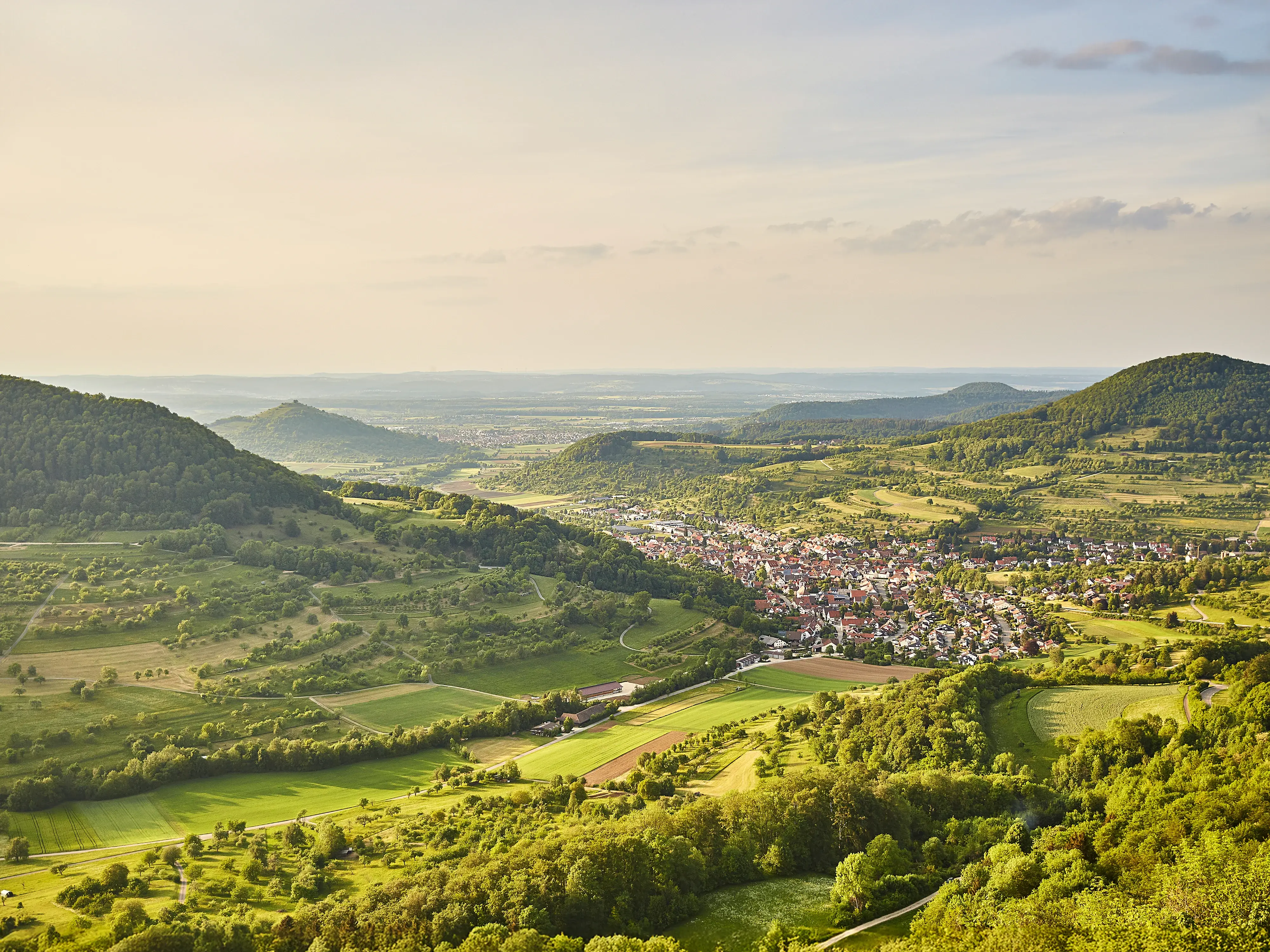Blick auf ein kleines Dorf in einem grünen, hügeligen Tal unter einem bewölkten Himmel.