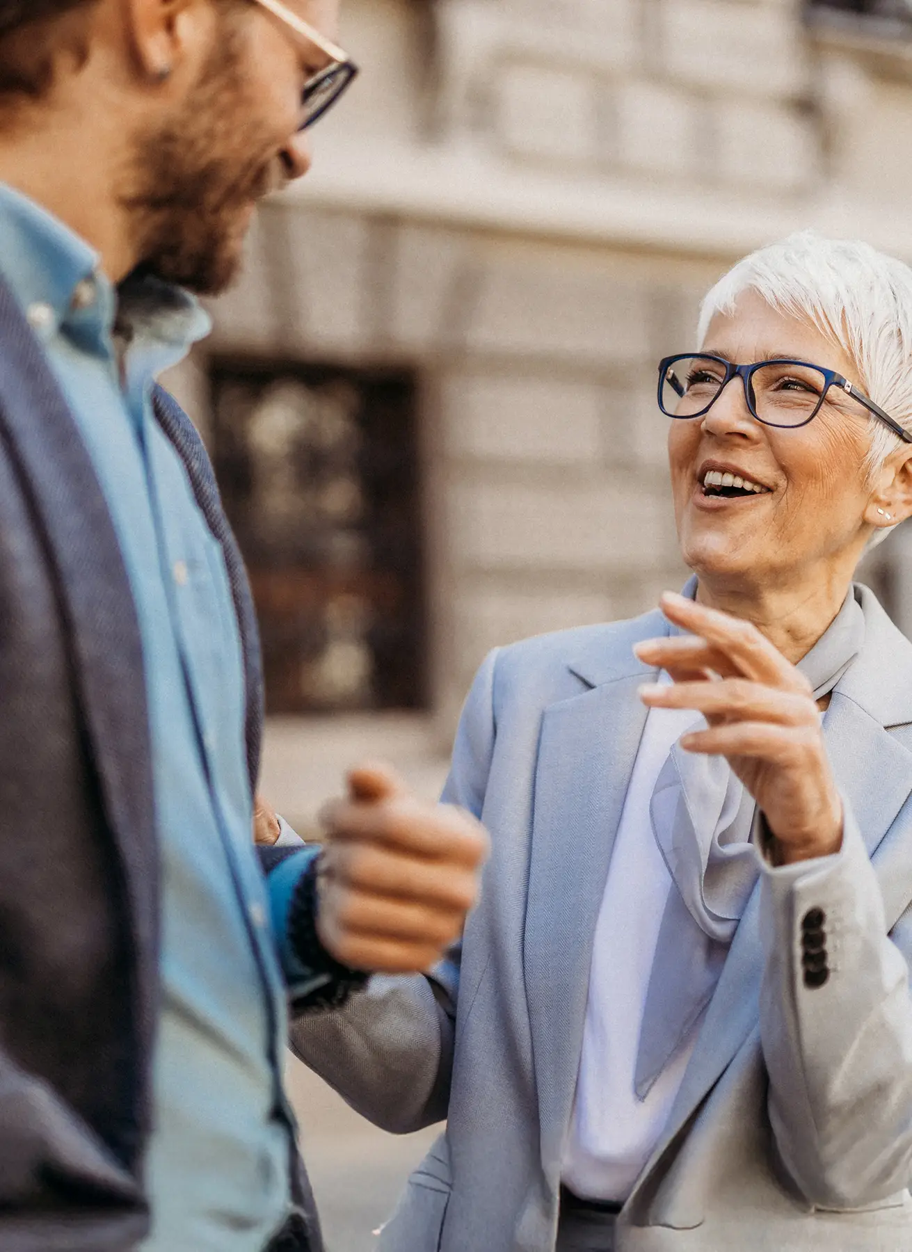 Ältere Frau mit kurzen weißen Haaren und blauer Brille im Gespräch mit einem Mann vor einem Gebäude.