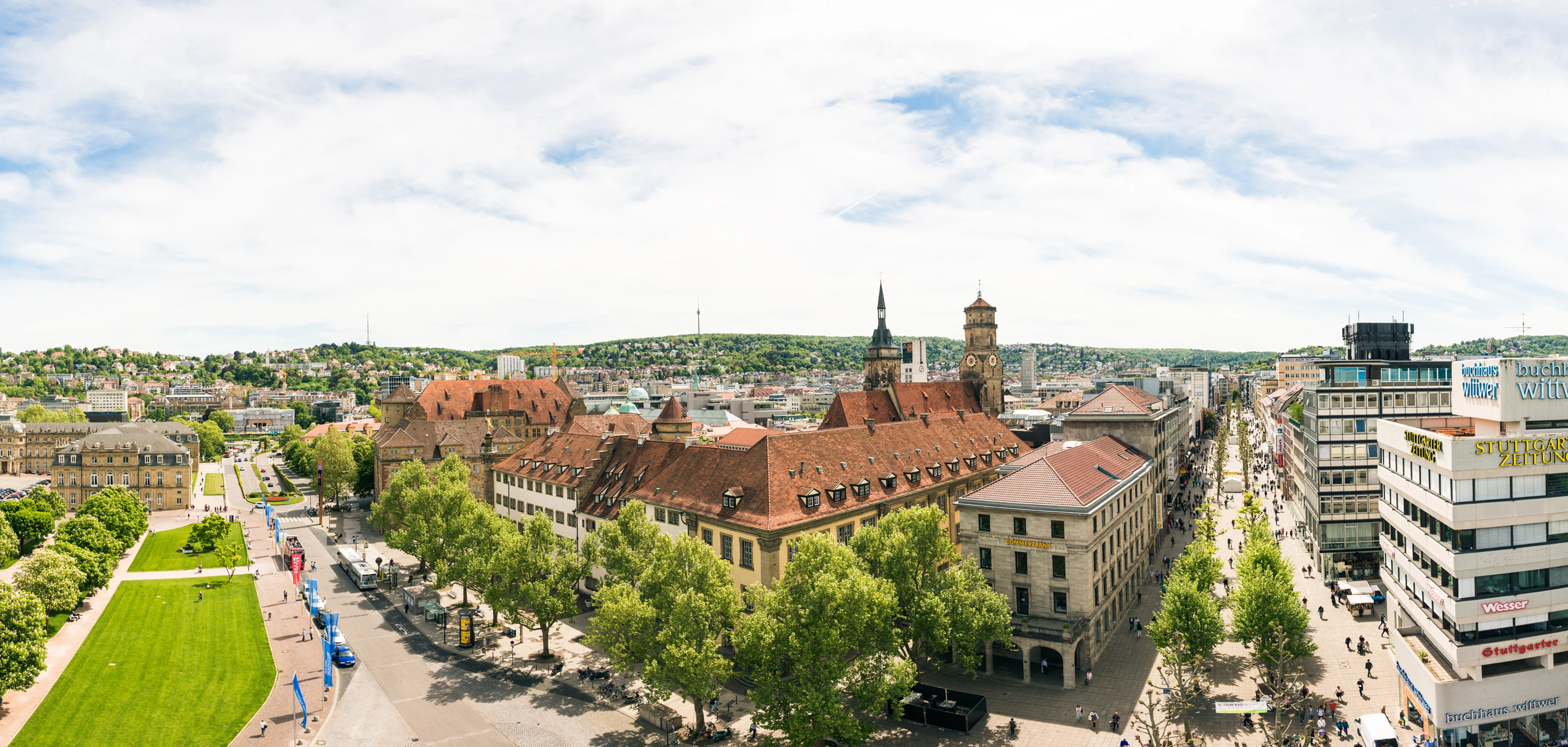 Panoramablick auf die Stuttgarter Innenstadt mit historischen Gebäuden, grünen Bäumen und einer belebten Einkaufsstraße an einem sonnigen Tag.