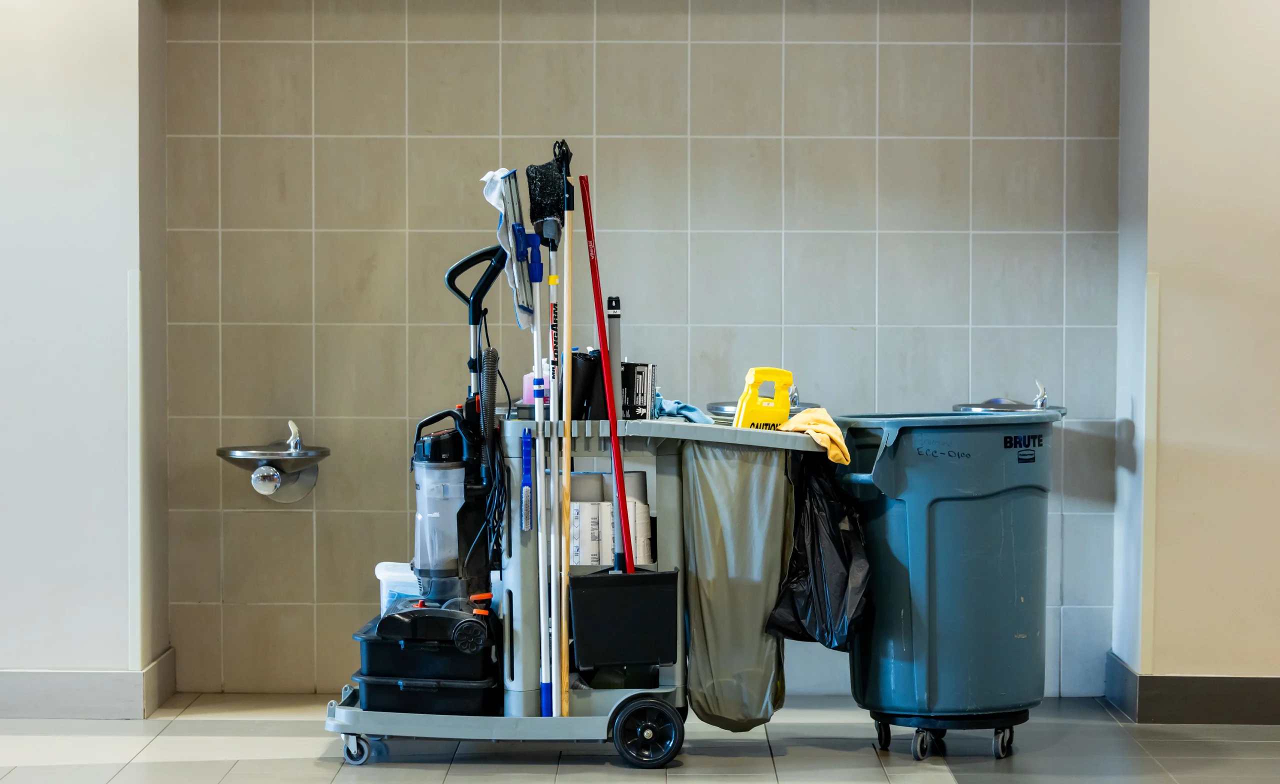 Janitorial cleaning cart with mop, vacuum, caution sign, trash bins, and cleaning supplies in a tiled hallway.