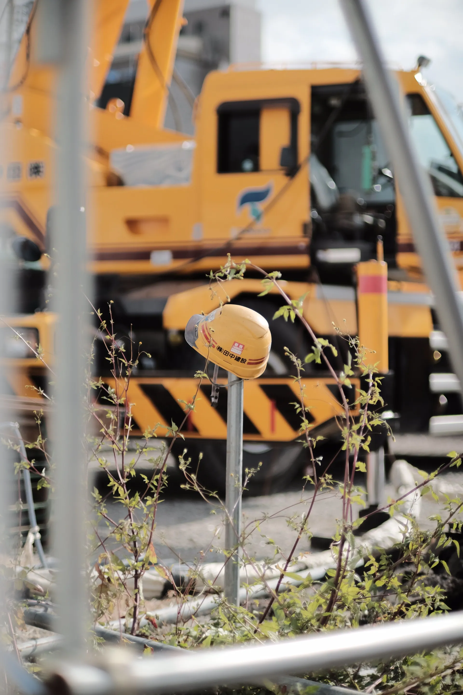 Yellow construction helmet on metal pole with yellow crane truck and plants in blurred background.