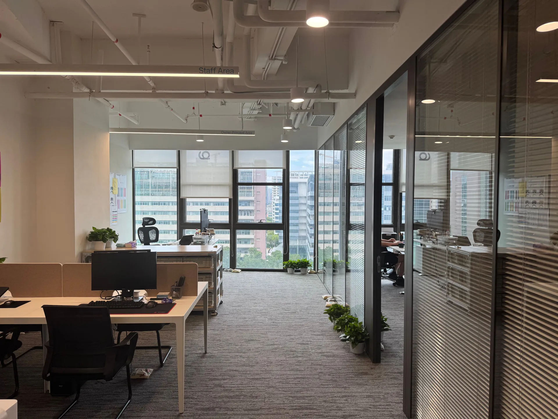 Modern office with workstations, black chairs, glass partitions, and large windows showing city buildings.
