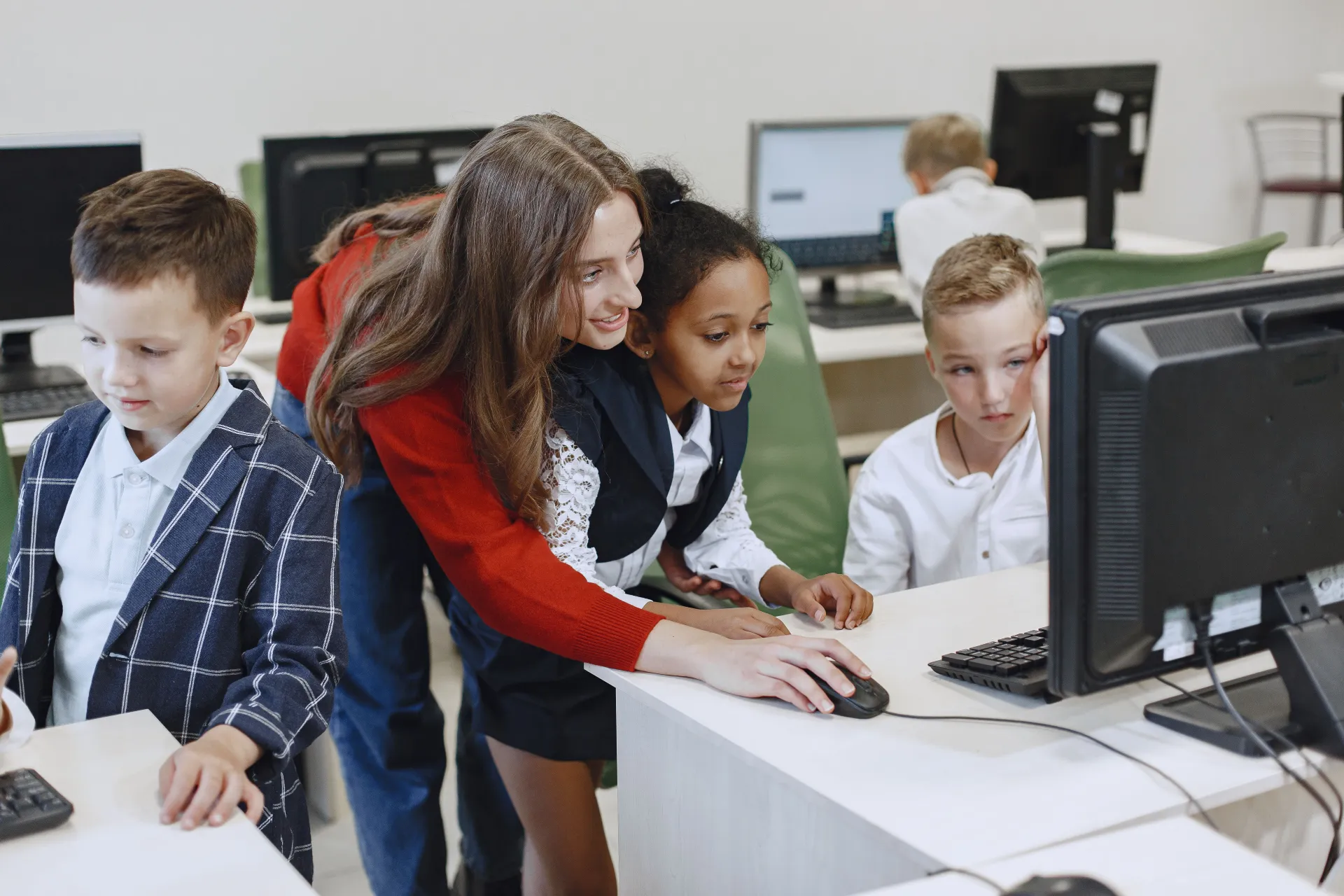 Adult woman teaches a group of children using computers in a classroom setting.