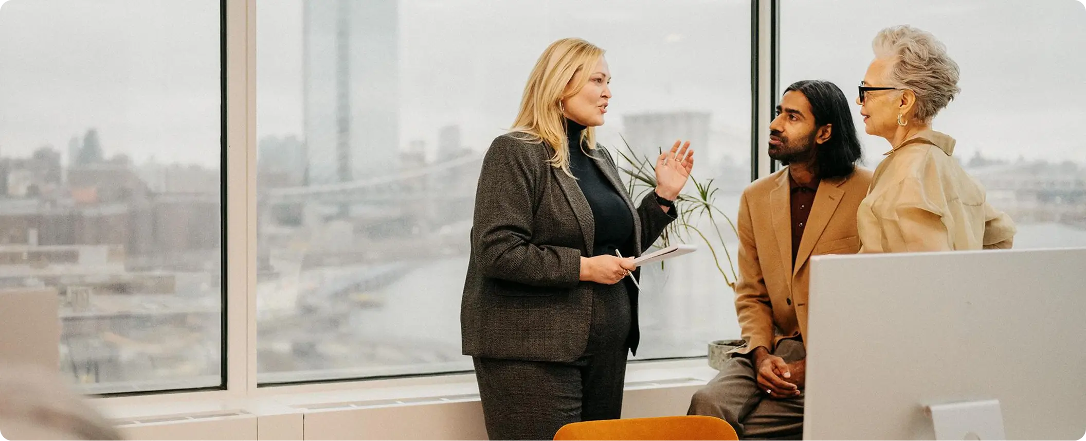 Three diverse colleagues engaged in a discussion in a modern office with large windows overlooking a cityscape.