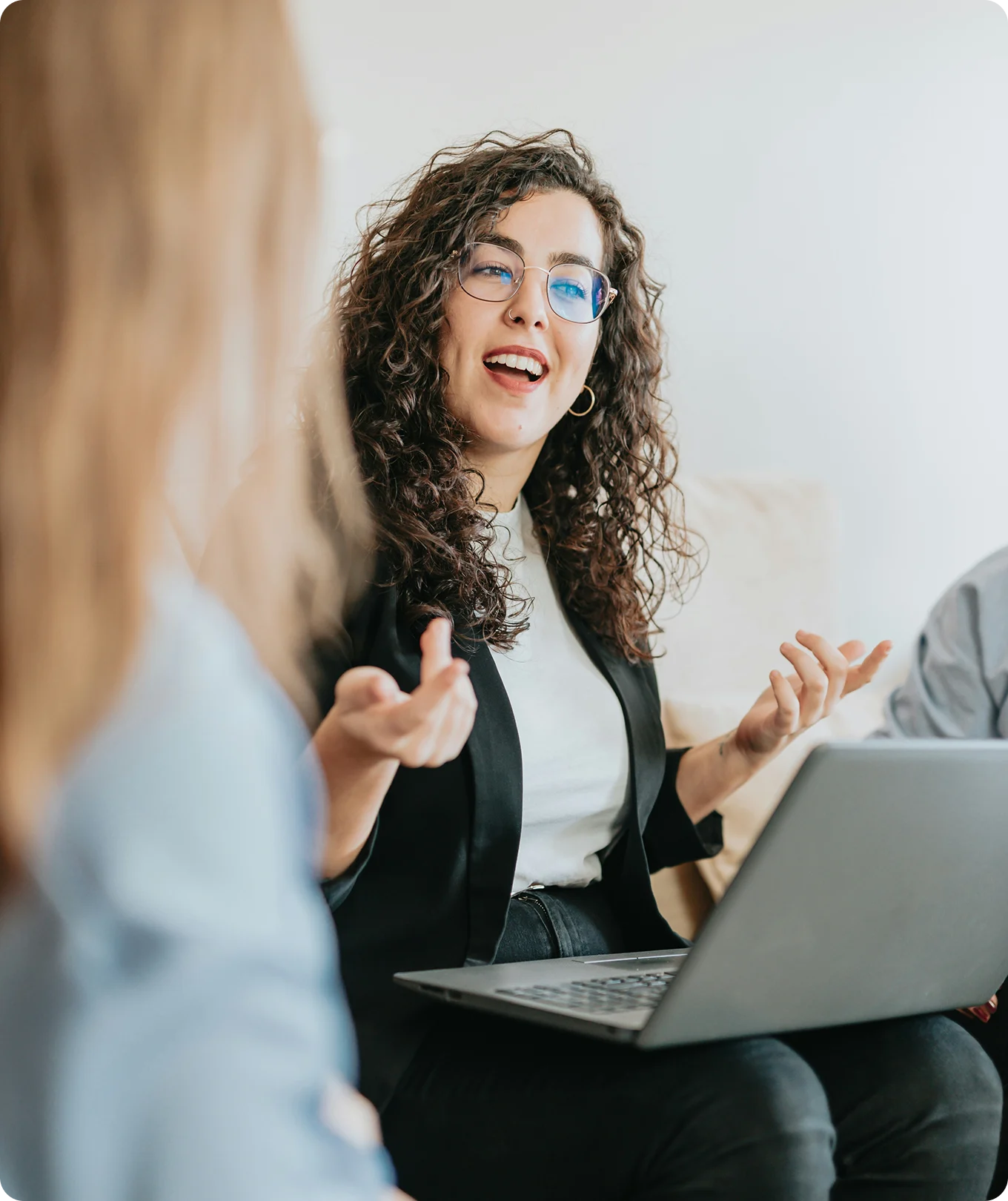 Curly-haired woman wearing glasses and a black blazer speaks animatedly while using a laptop.