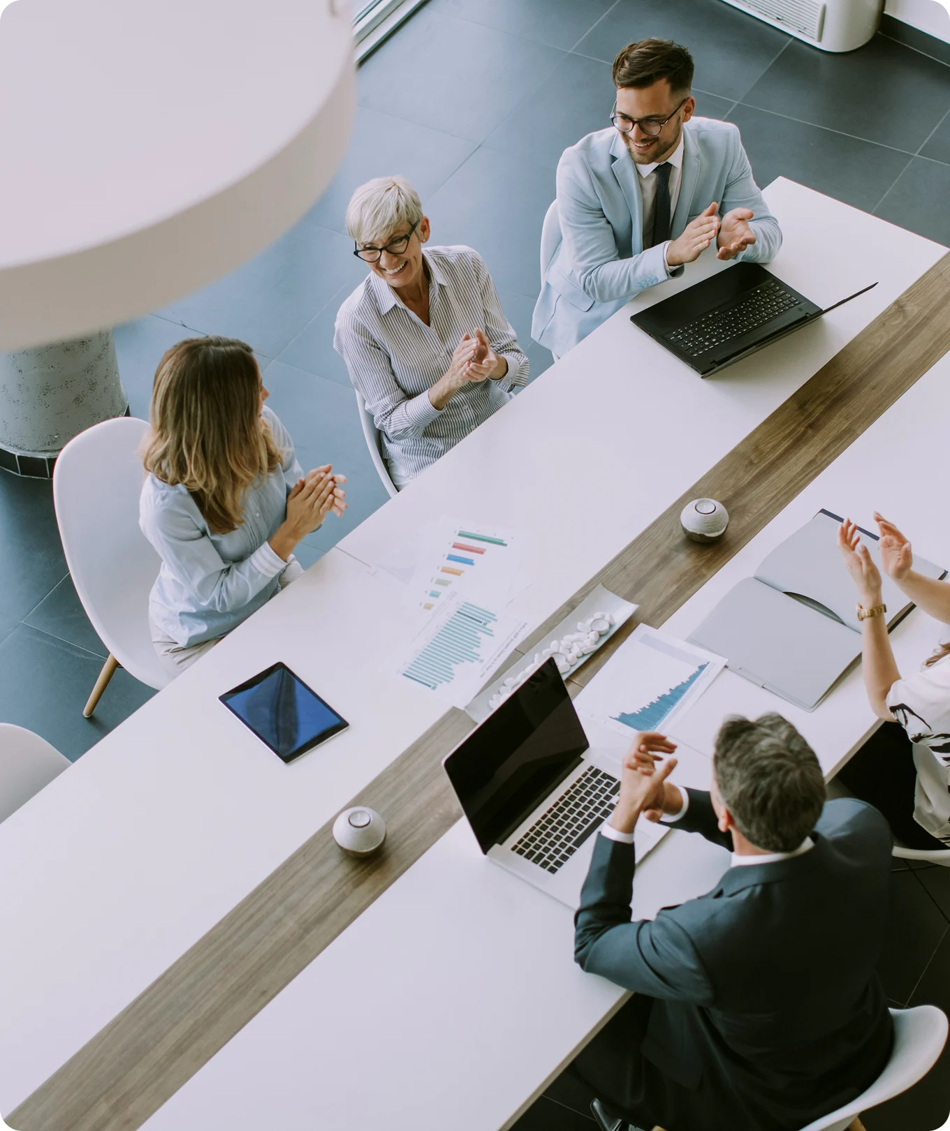 Four business professionals sitting around a table clapping during a meeting with laptops and charts on the table.