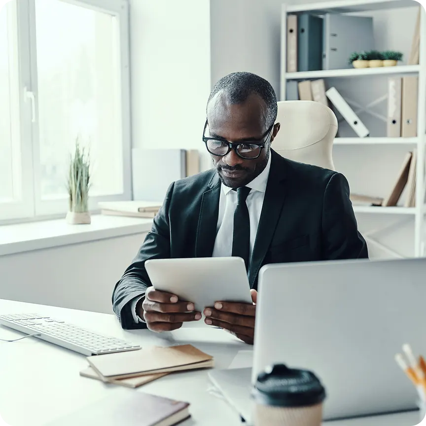 Man in glasses and suit sitting at a desk using a tablet, with a laptop, keyboard, notebooks, and coffee cup nearby in a bright office.