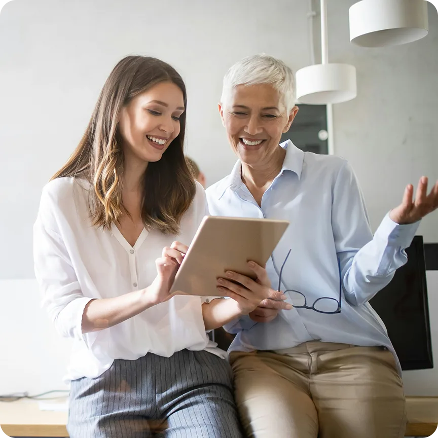 Two women smiling and looking at a tablet screen together in a bright office setting.